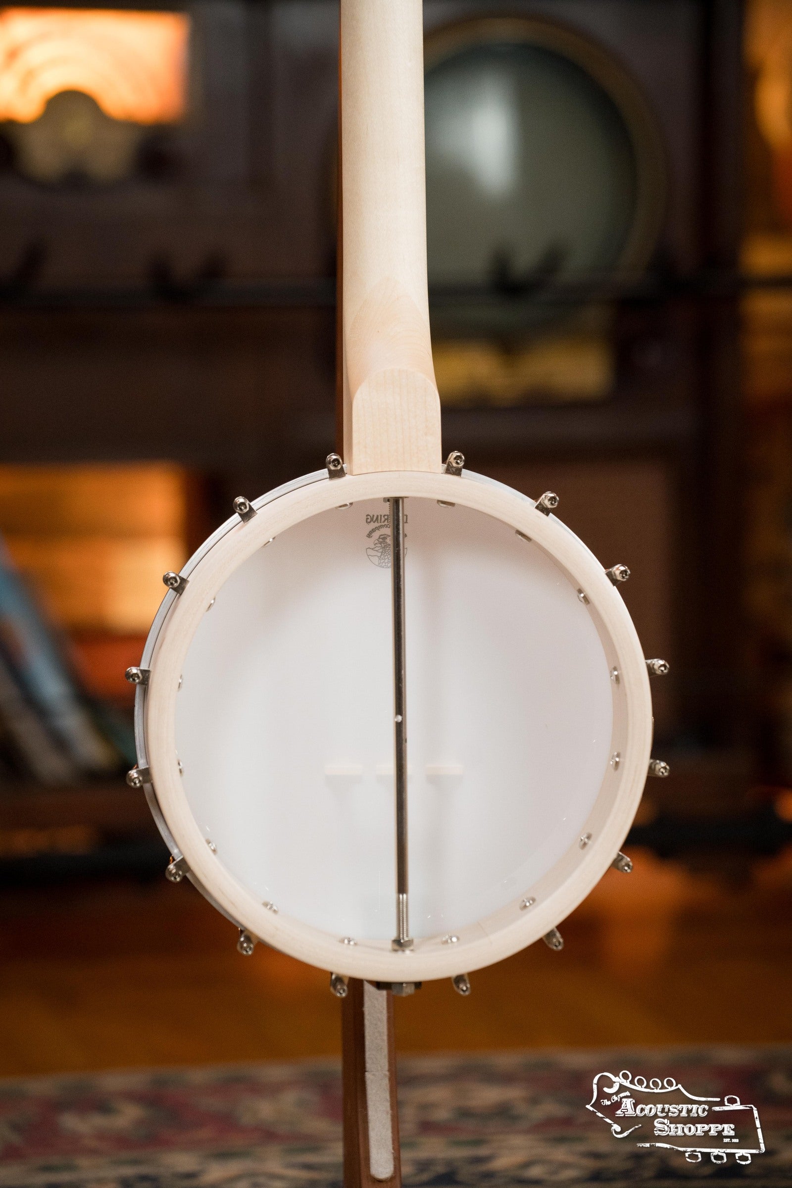 A close-up of the Deering Banjos Goodtime Deco 5-String Banjo #52407 shows its round body and strings facing forward, with shelves and musical gear blurred in the background. The Acoustic Shoppe logo appears at the bottom right.