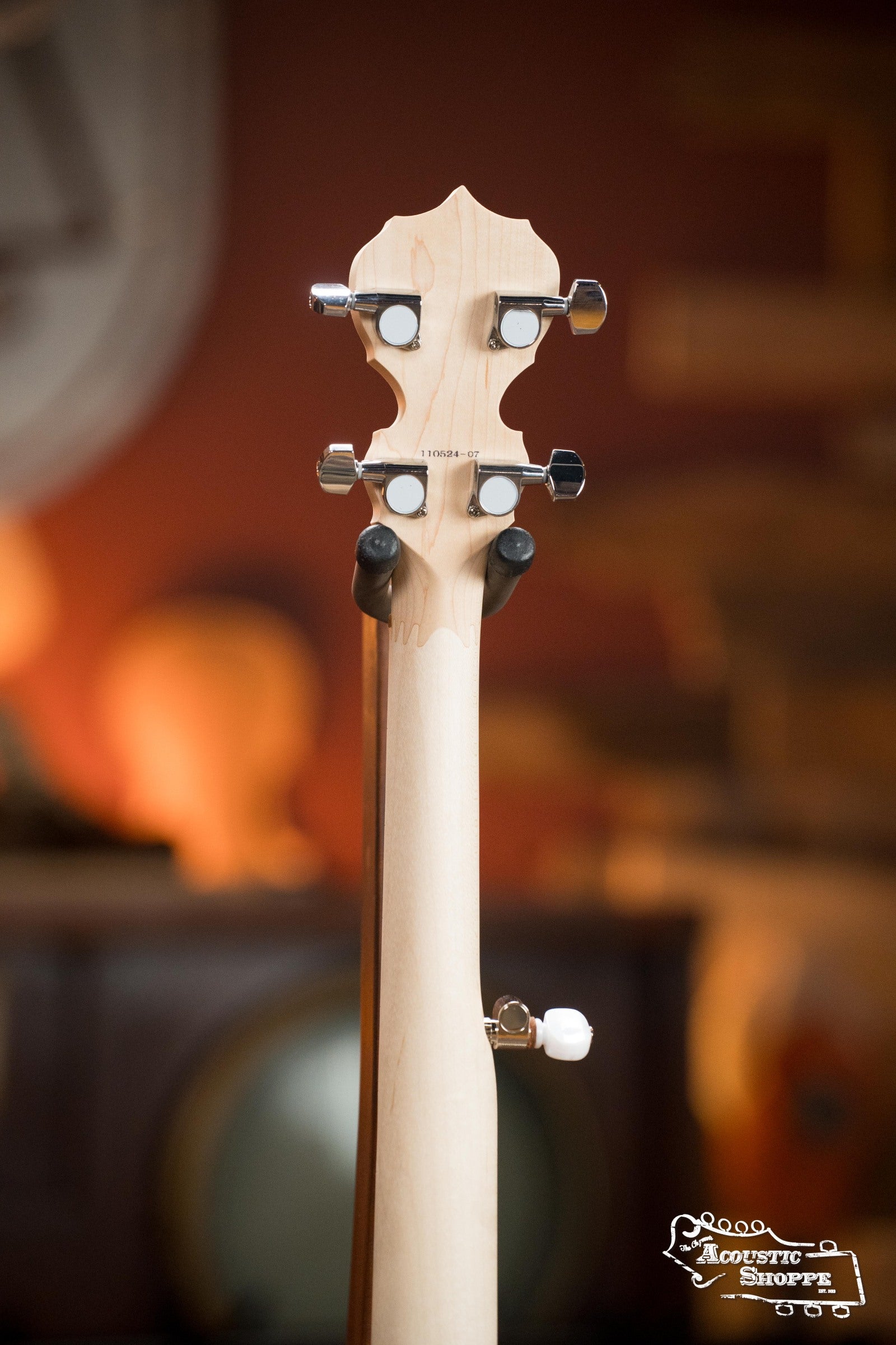 A close-up of the Deering Goodtime Deco 5-String Banjo #52407 headstock by Deering Banjos shows four tuning pegs and a light-colored neck, set against a softly blurred, warm-toned vintage background.