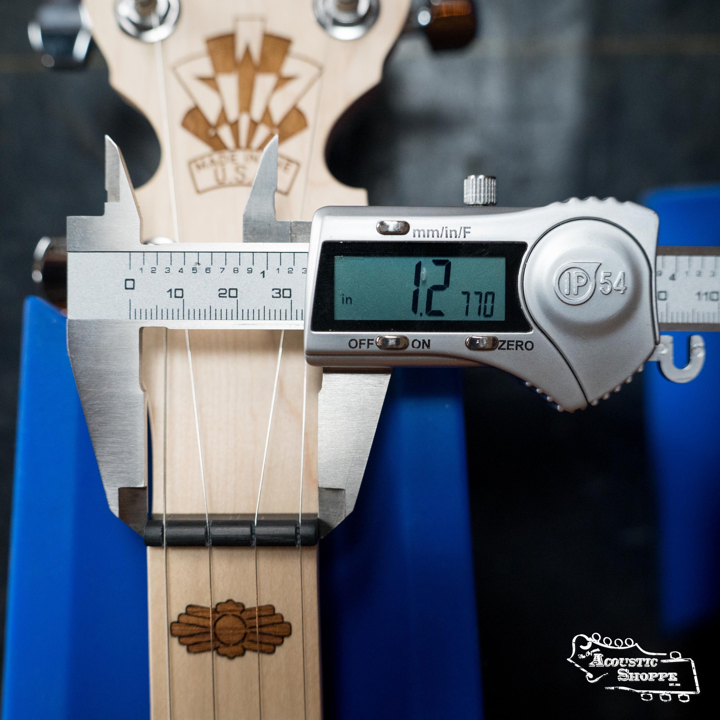 A digital caliper measures the Deering Goodtime Deco 5-String Banjo #52407 neck at 1.277 inches wide. The Deering Banjos instrument rests on a blue support, with the Acoustic Shoppe logo shown in the bottom right corner.