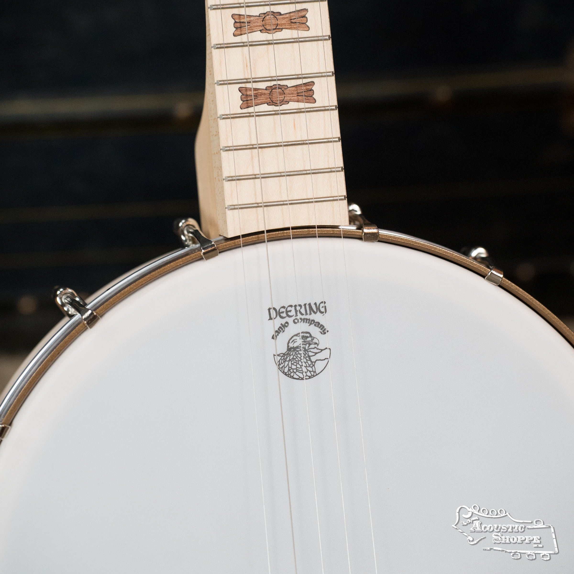 Close-up of the Deering Goodtime Deco 5-String Banjo #62407 by Deering Banjos, highlighting its wooden neck with Art Deco inlays and a white drumhead bearing the Deering logo against a dark, blurred background.