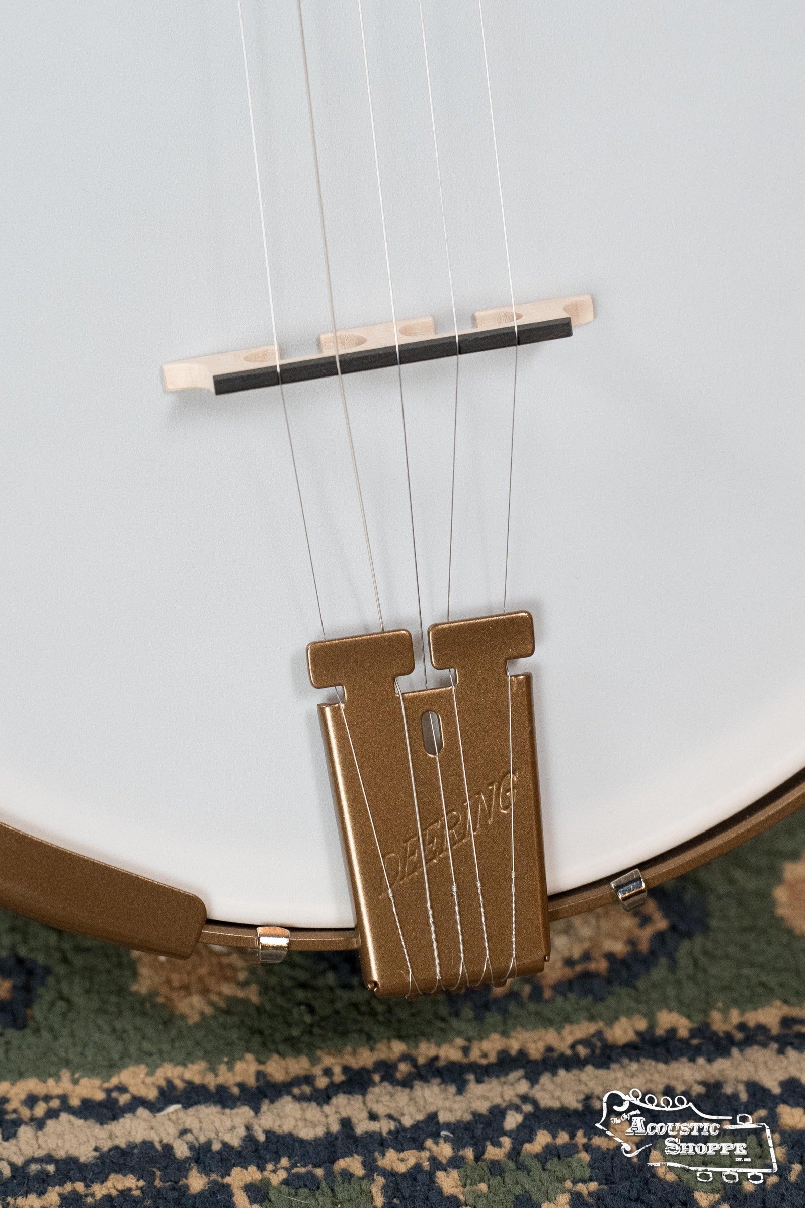 Close-up of a Deering Banjos Goodtime Deco 5-String Banjo #62407 bridge and tailpiece on patterned carpet; DEERING is engraved on the tailpiece, with the Acoustic Shoppe logo in the lower right corner.