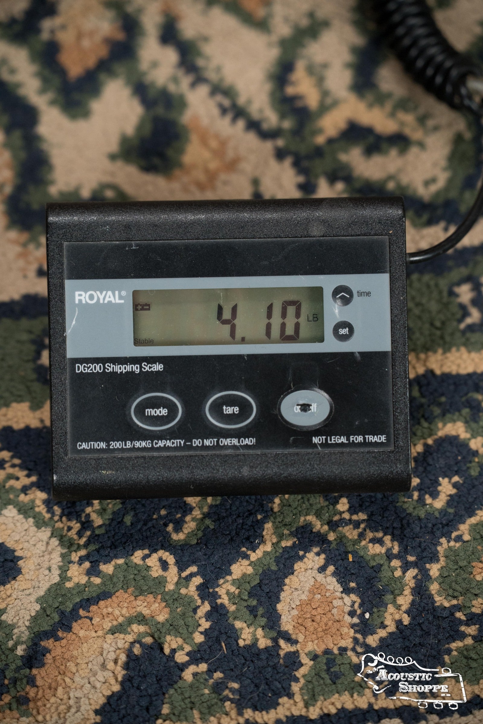 A Royal DG200 Shipping Scale on a patterned rug shows 4.10 pounds—the weight of a Deering Goodtime Deco 5-String Banjo #62407 by Deering Banjos. The black scale has gray buttons and Royal DG200 Shipping Scale printed on it.