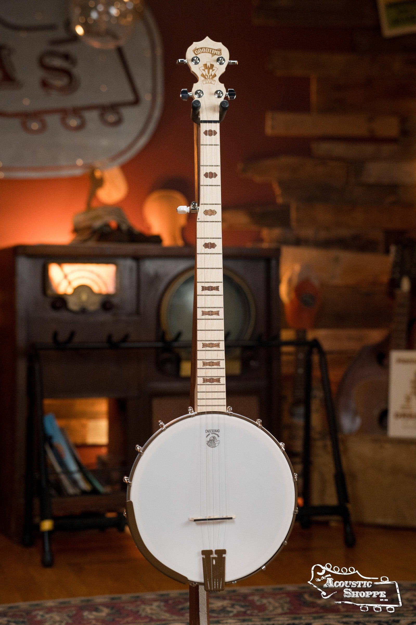 A Deering Banjos Goodtime Deco 5-String Banjo #62407 stands upright on a stand in a warmly lit room with wooden walls, vintage decor, and the Acoustic Shoppe logo in the bottom right corner.