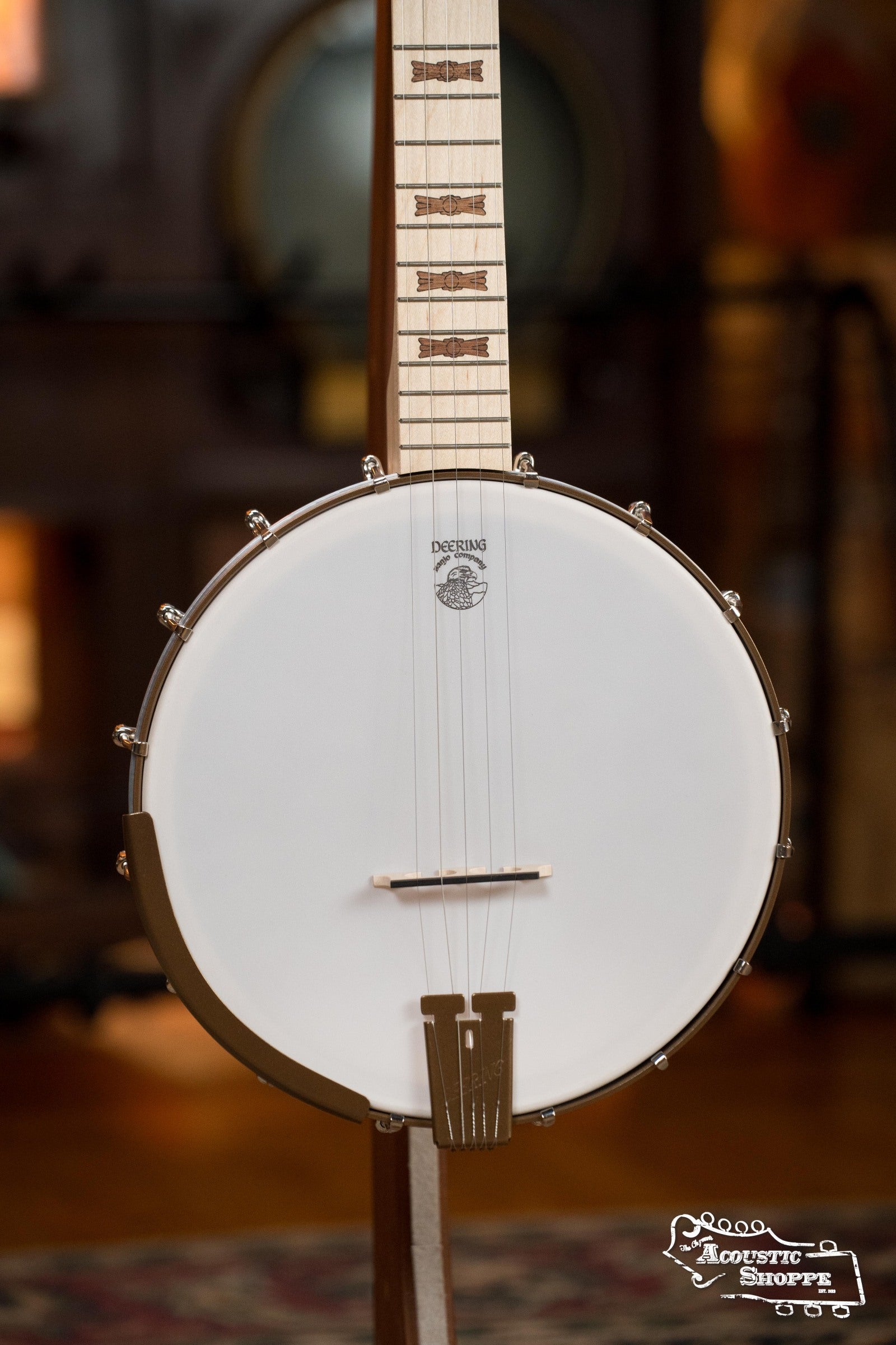 A Deering Banjos Goodtime Deco 5-String Banjo #62407 with a light wood neck and white drumhead is displayed upright indoors, featuring The Acoustic Shoppe logo in the bottom right corner.