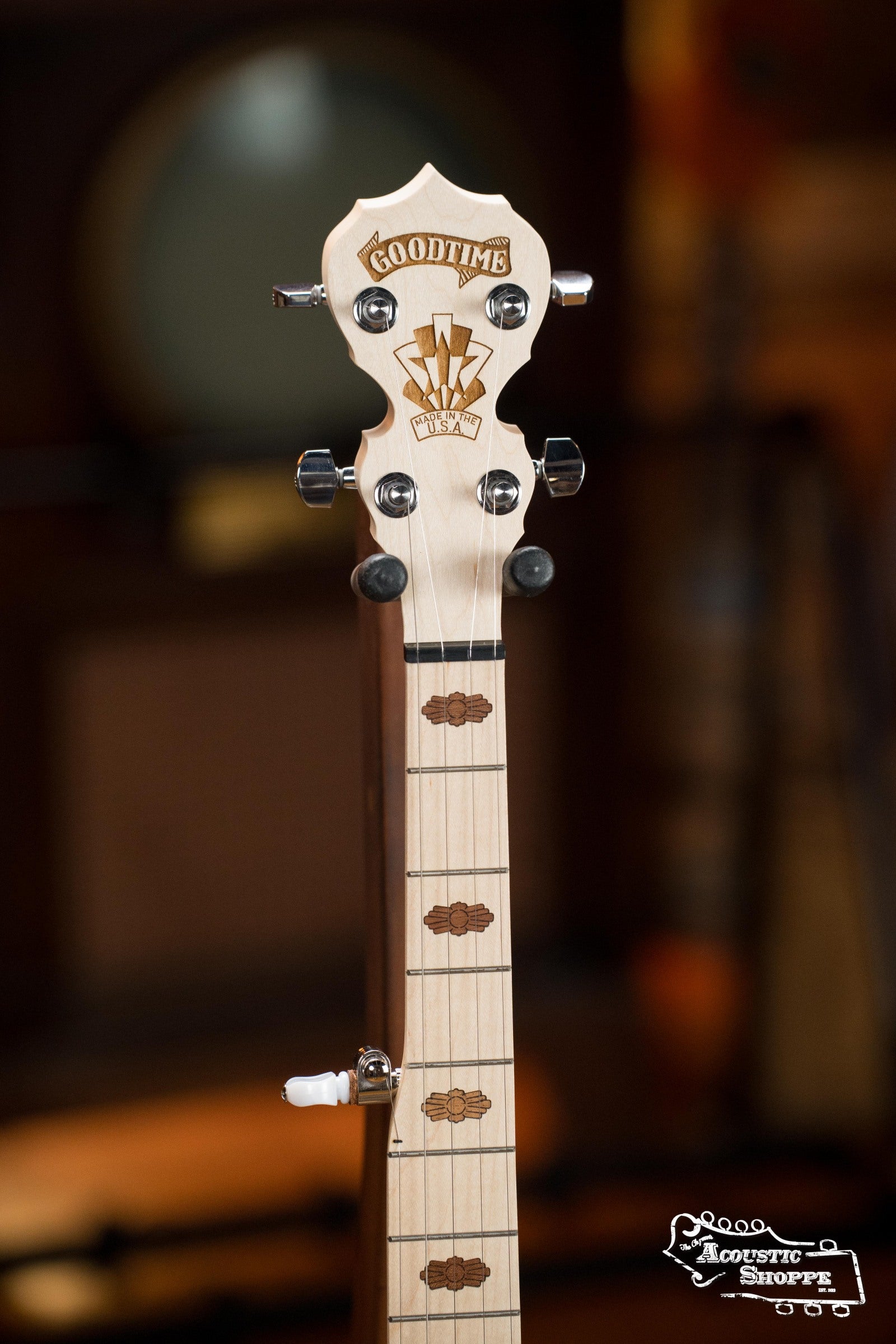 Close-up of the headstock and upper neck of a Deering Banjos Goodtime Deco 5-String Banjo #62407, featuring decorative inlays and tuning pegs. The Acoustic Shoppe logo appears bottom right, with a blurred background highlighting the design.