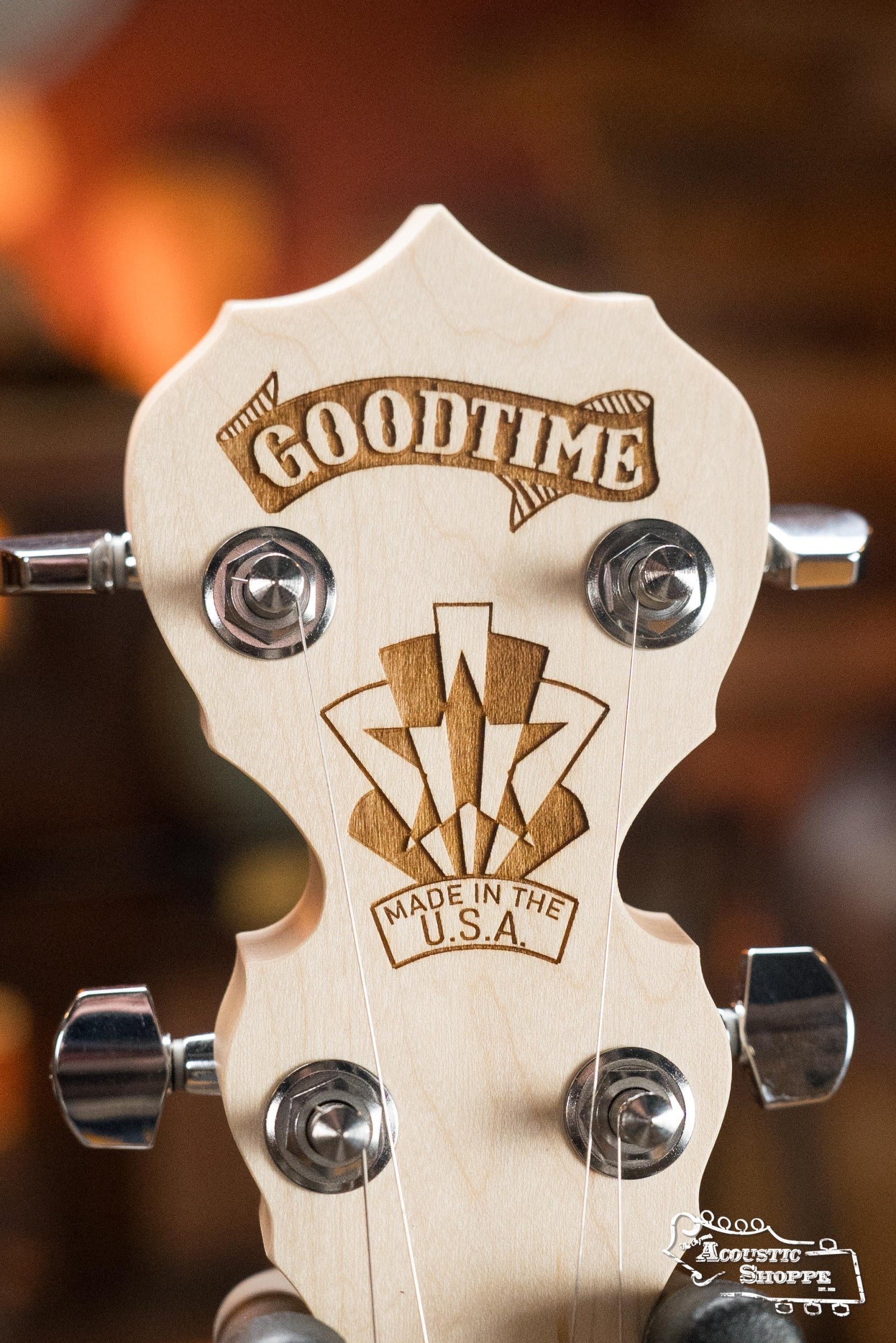 Close-up of a Deering Banjos Goodtime Deco 5-String Banjo #62407 headstock showing GOODTIME and MADE IN THE U.S.A. engravings with metal tuners, set against a warmly blurred background.