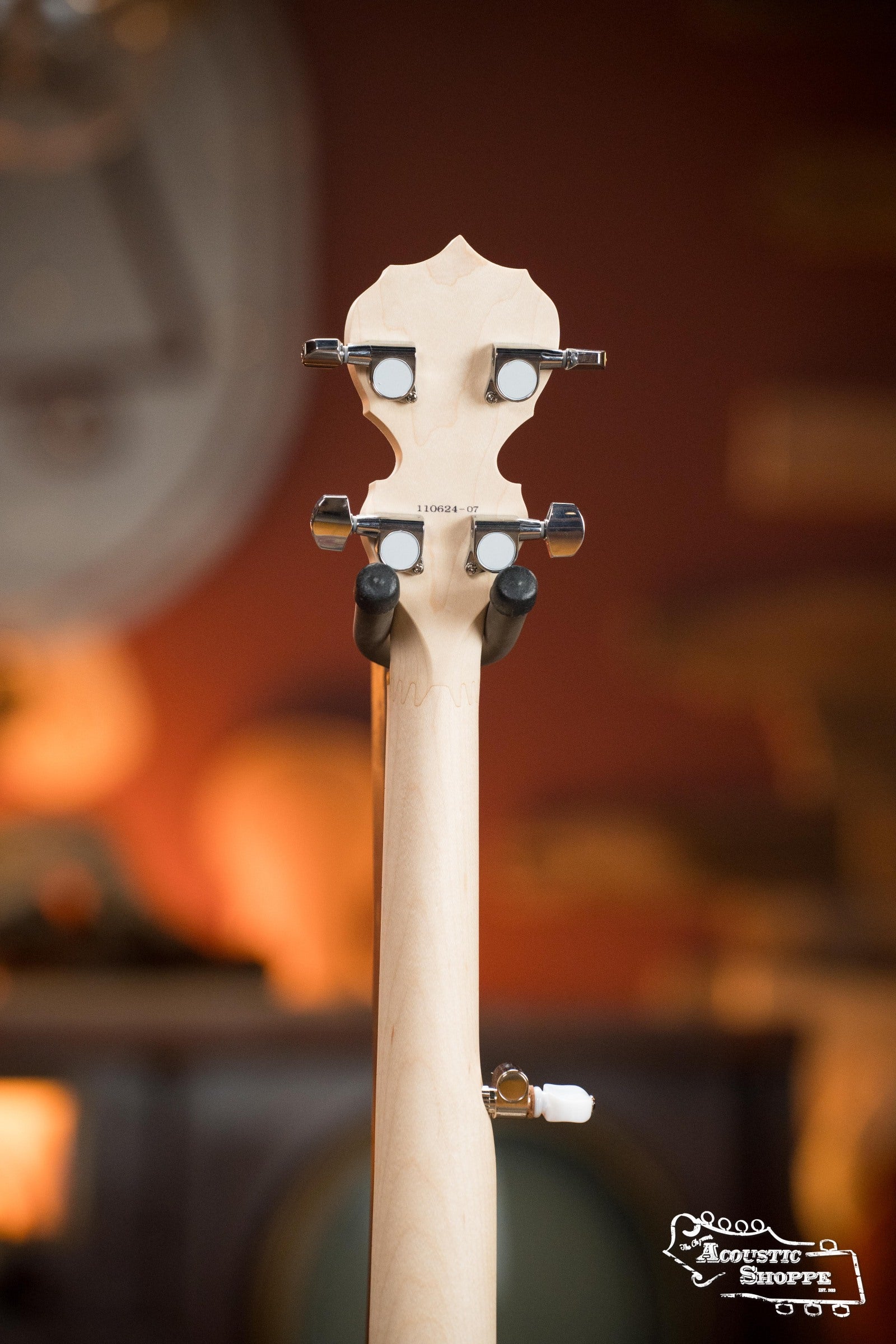 The back of a light-colored Deering Banjos Goodtime Deco 5-String Banjo #62407 headstock with tuning pegs is shown against a warm, blurred background, with the Acoustic Shoppe logo in the bottom right corner.
