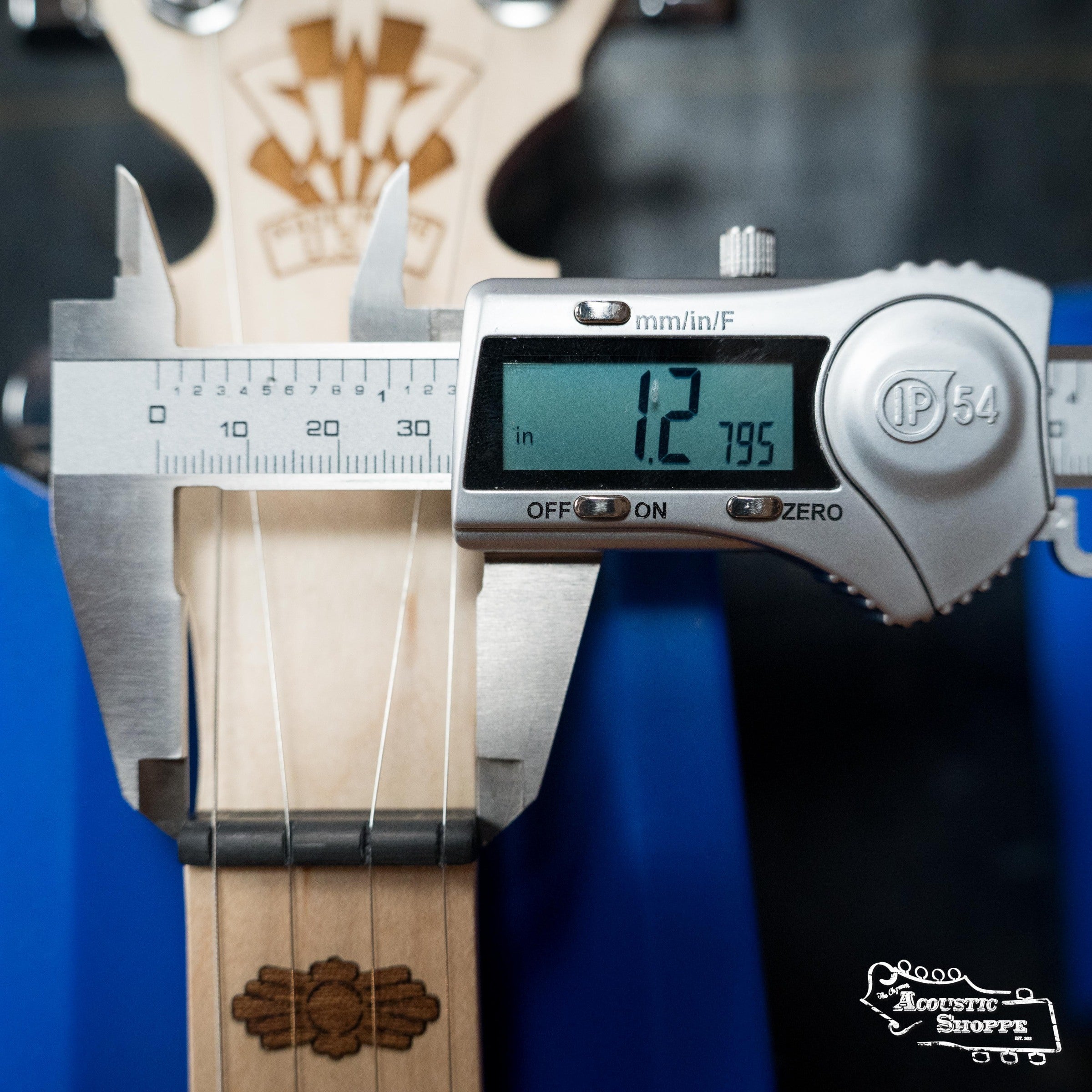 A caliper measures the Deering Goodtime Deco 5-String Banjo #62407 neck at 1.2 inches near the headstock, next to Art Deco-style inlays; the reading is shown on a digital screen. Brand: Deering Banjos.