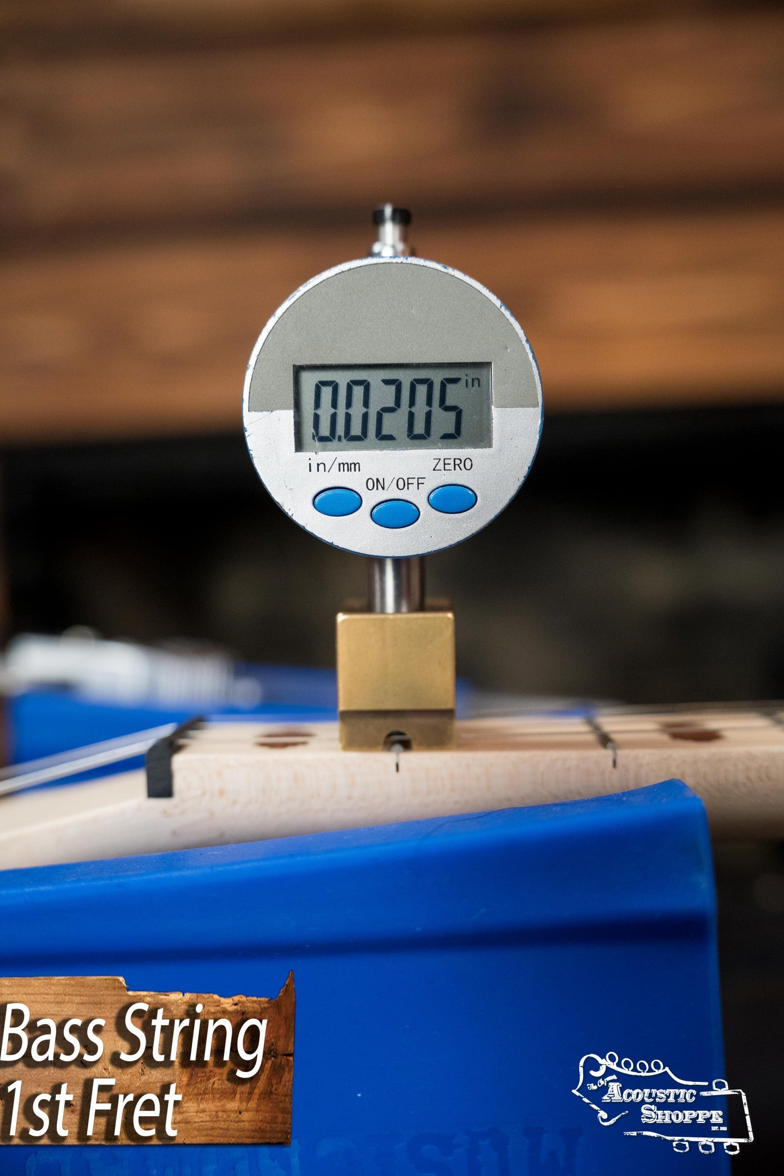 A digital gauge displays a 0.0205-inch bass string height at the 1st fret of a Deering Goodtime Deco 5-String Banjo #62407 by Deering Banjos, with a blue background and “Bass String 1st Fret” text in the lower left.