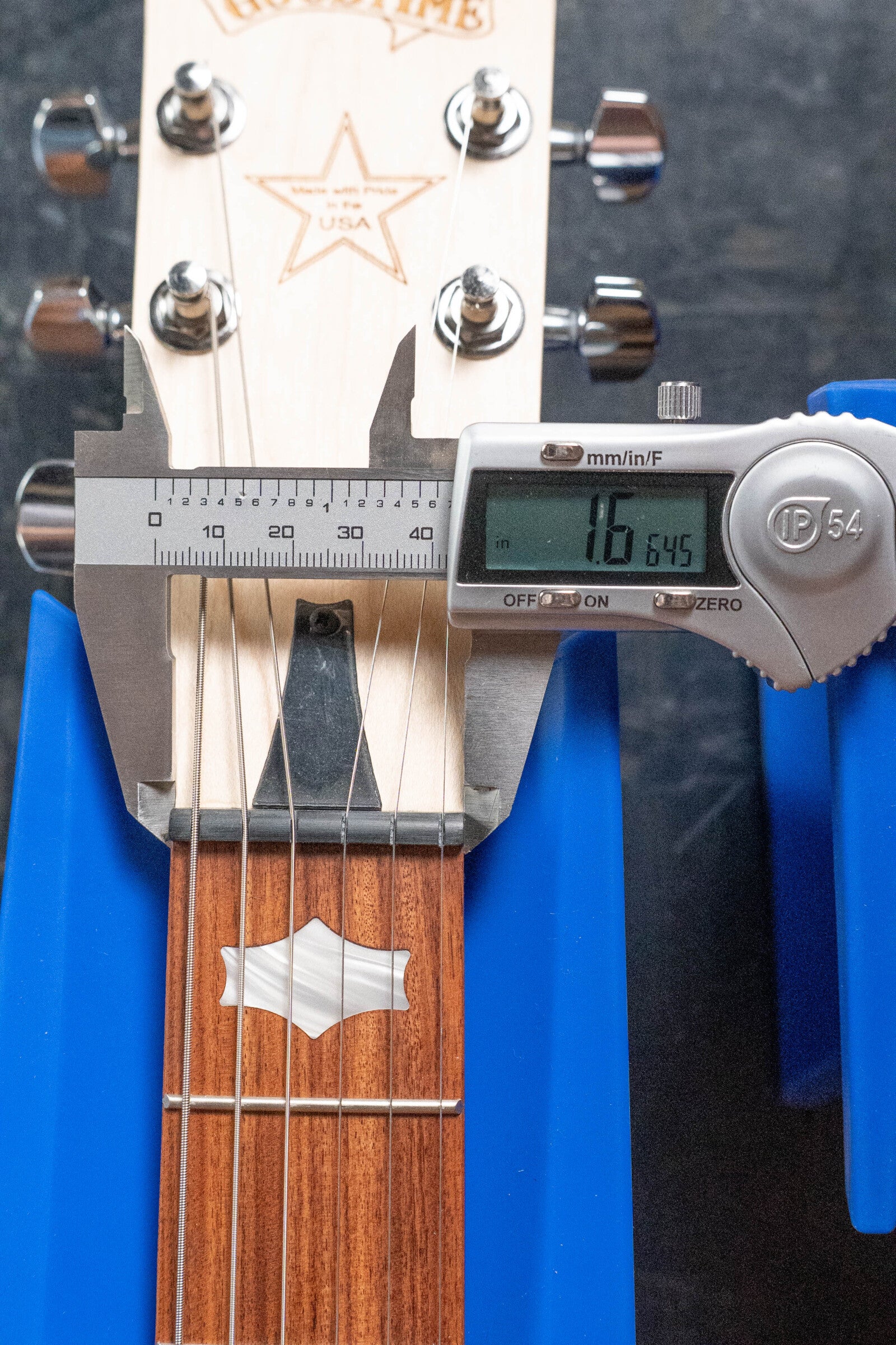 A Deering Goodtime Six-R 6-String Resonator Banjo w/Kavanjo Pickup by Deering Banjos is measured with a digital caliper at 1.645 inches neck width, shown on blue supports, featuring metal tuning pegs and a fretboard inlay.