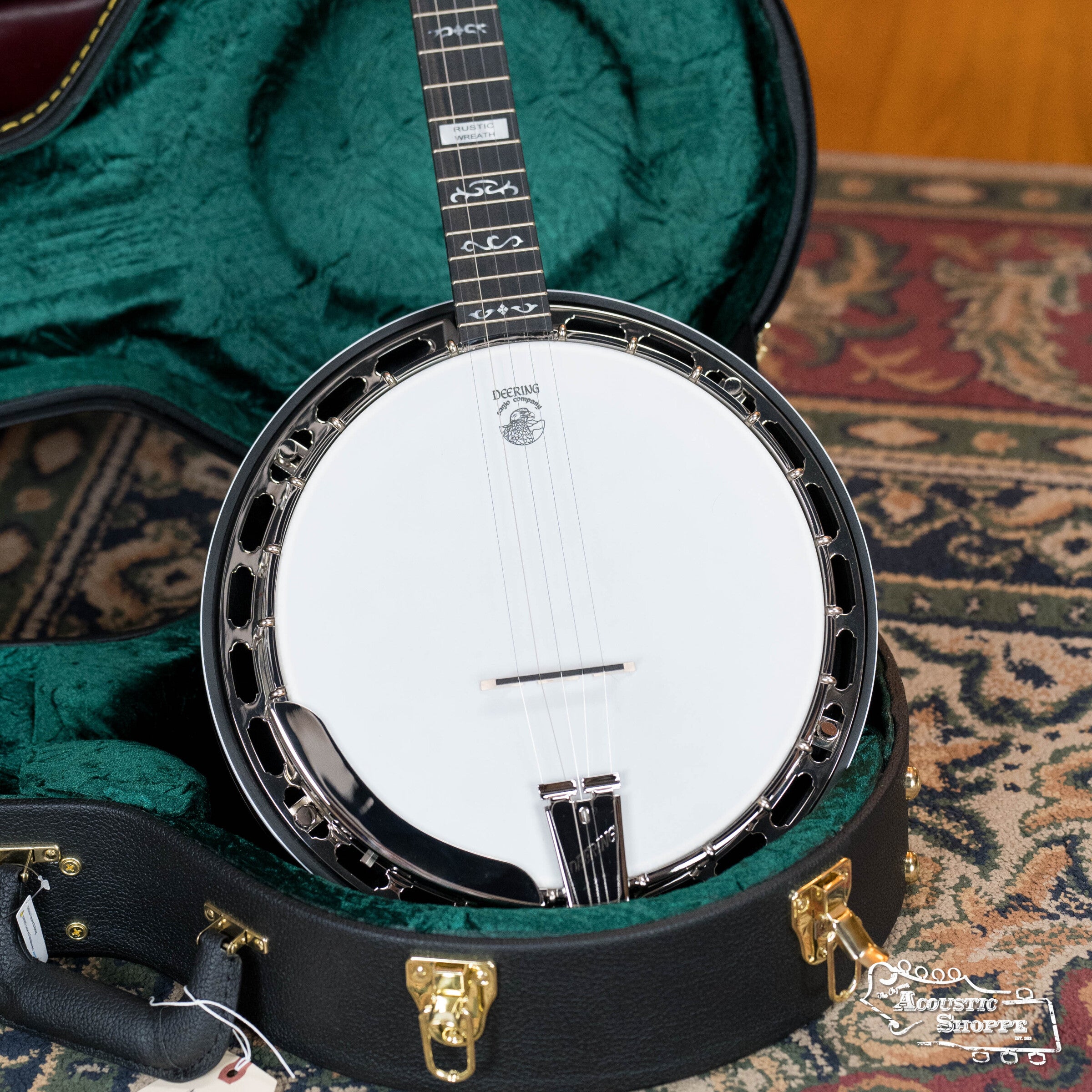 A Deering Rustic Wreath 5-String Resonator Banjo #AU45 by Deering Banjos, featuring a white drumhead, black neck with pearl inlays, rests in a green-lined hard case showing gold latches and handle on a patterned carpet.