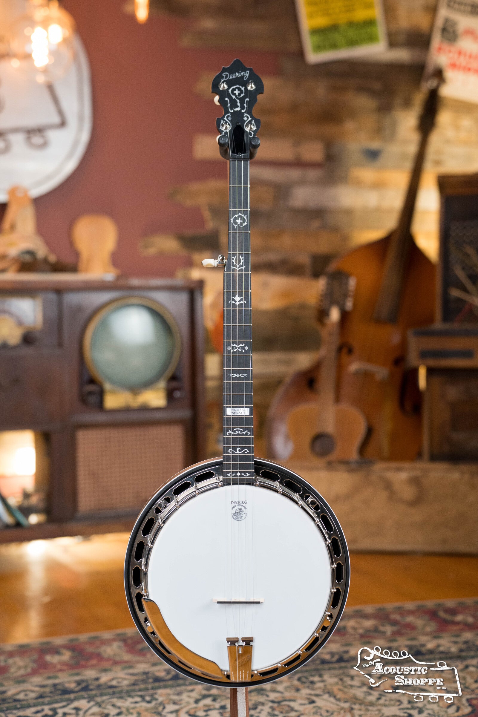 A Deering Rustic Wreath 5-String Resonator Banjo #AU45 by Deering Banjos with a black headstock stands upright in a vintage room with wooden walls, a double bass, retro TV, patterned rug, and warm lighting.