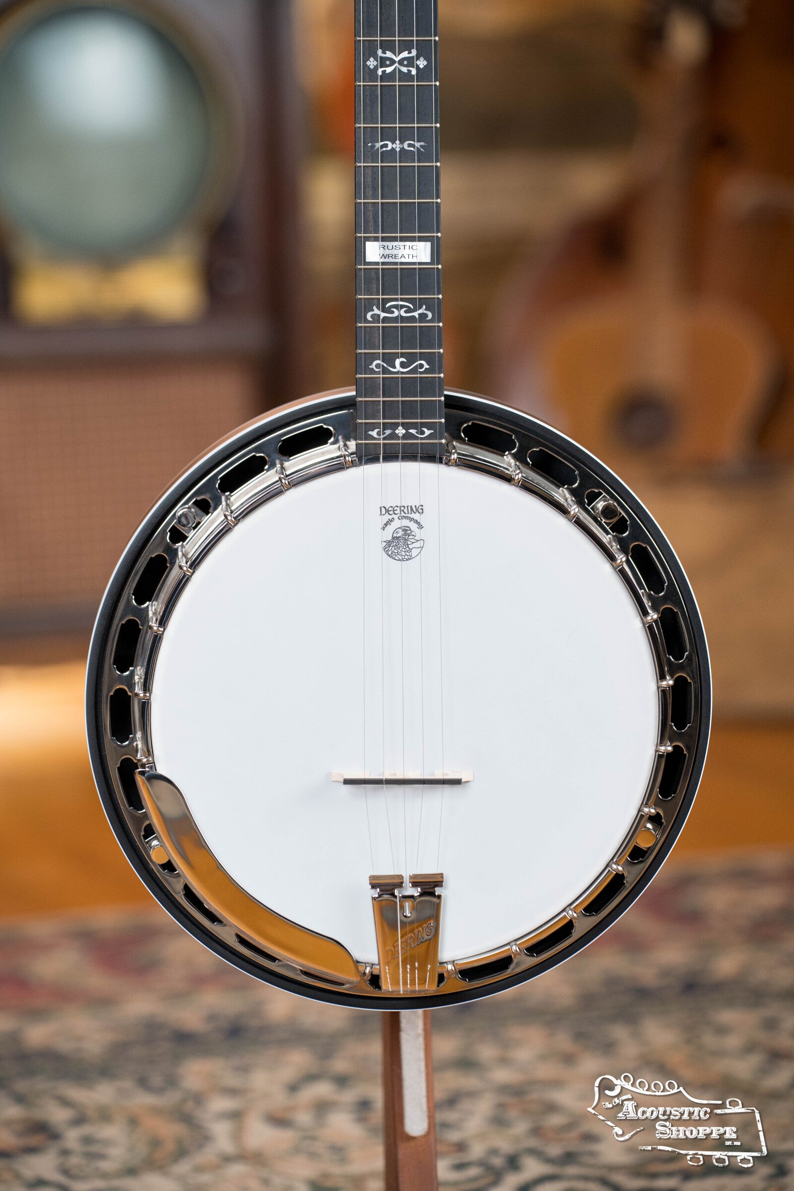 A close-up of the Deering Banjos Rustic Wreath 5-String Resonator Banjo #AU45, with a white drumhead, metal rim, and decorative neck inlays, stands upright on a patterned rug amid blurred musical instruments.