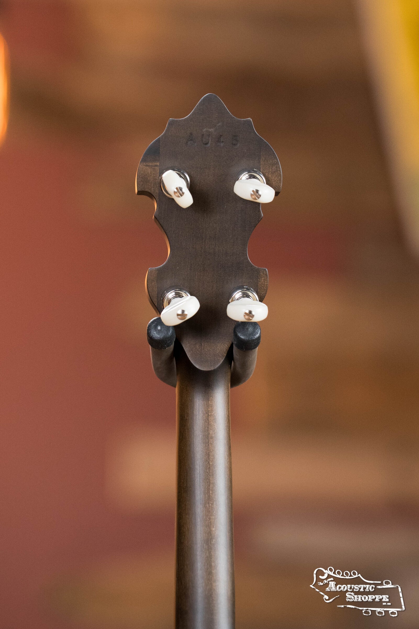 The back of a Deering Banjos Rustic Wreath 5-String Resonator Banjo #AU45 headstock with four tuning pegs rests on a stand, highlighting its classic design. Acoustic Shoppe logo is seen in the bottom right corner.