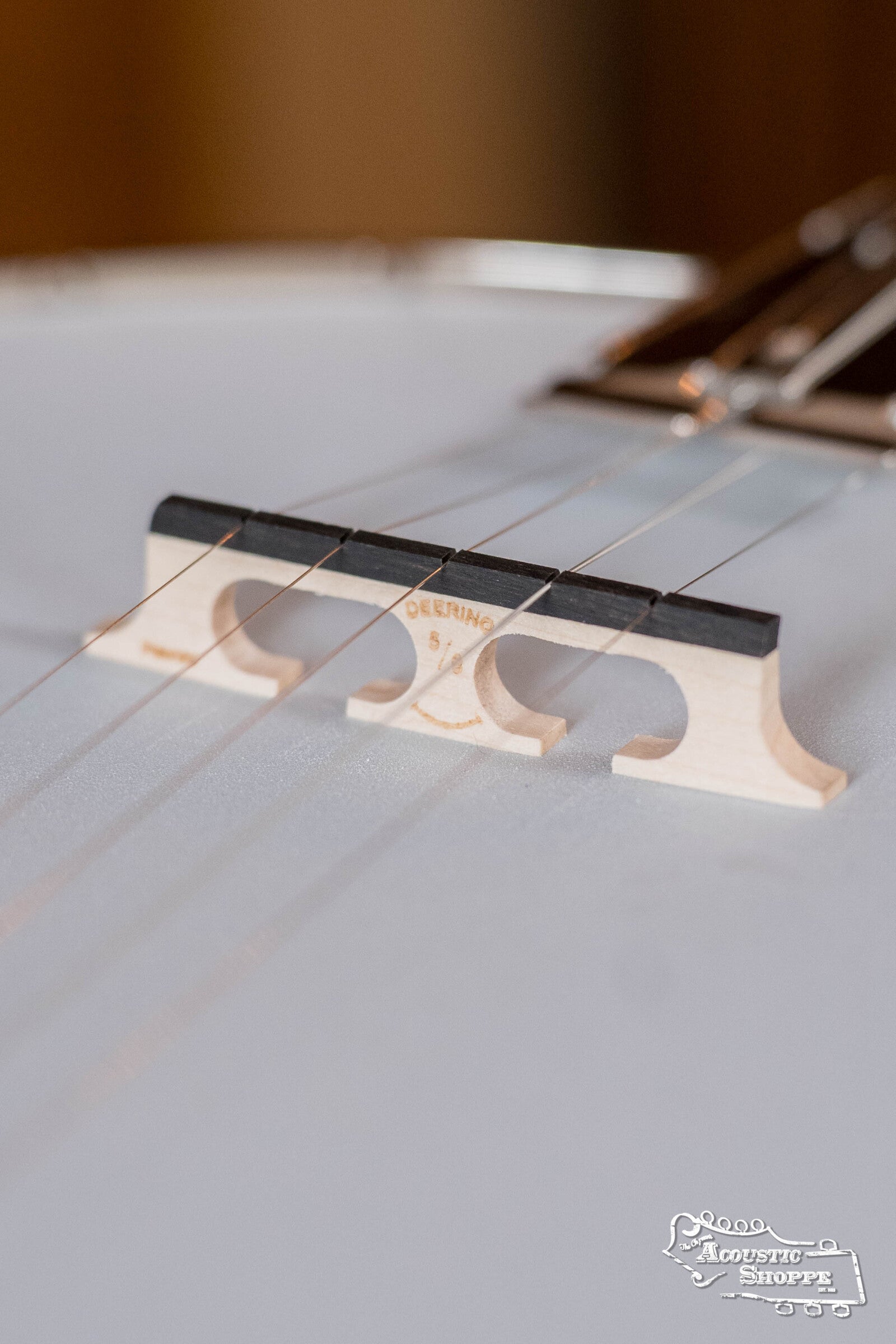 Close-up of a Deering Banjos Rustic Wreath 5-String Resonator Banjo #AU45 bridge with metal strings above it. The wooden bridge, marked “Deering,” is in sharp focus, with a blurred background and a logo at the bottom right corner.