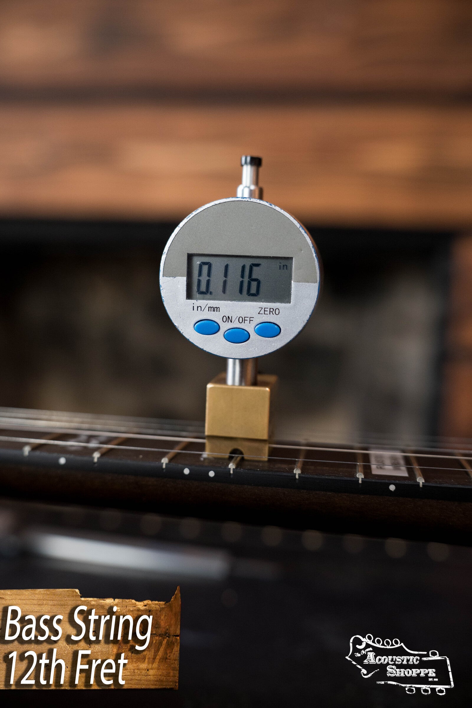 A digital gauge measures string height at the 12th fret of the Deering Rustic Wreath 5-String Resonator Banjo #AU45 by Deering Banjos, displaying 0.116 in., with a blurred background and The Acoustic Shoppe logo—ideal for Bluegrass Banjo fans seeking precision.