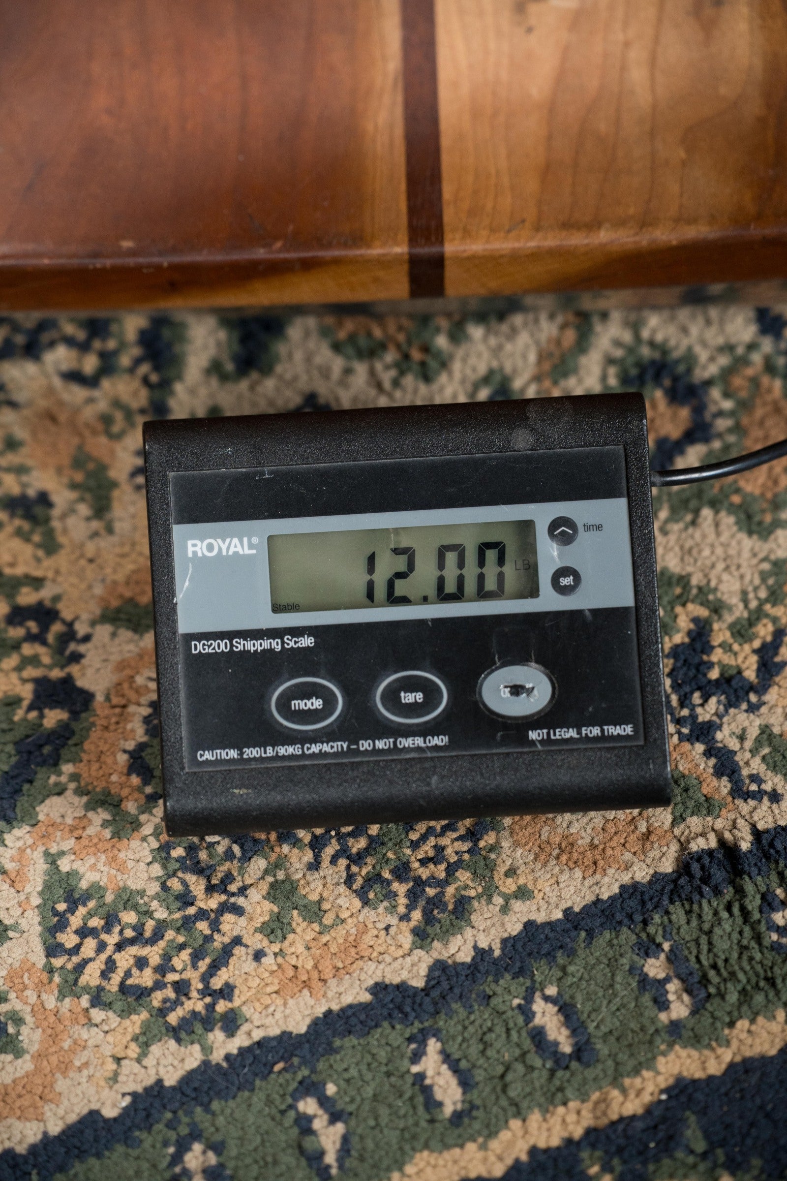 A digital shipping scale on a patterned carpet shows 12.00 lbs—the exact weight of the Deering Banjos Tony Trischka Silver Clipper Resonator Banjo w/Spikes at 7, 8, 9, and 10 (#0100)—ideal for bluegrass players.