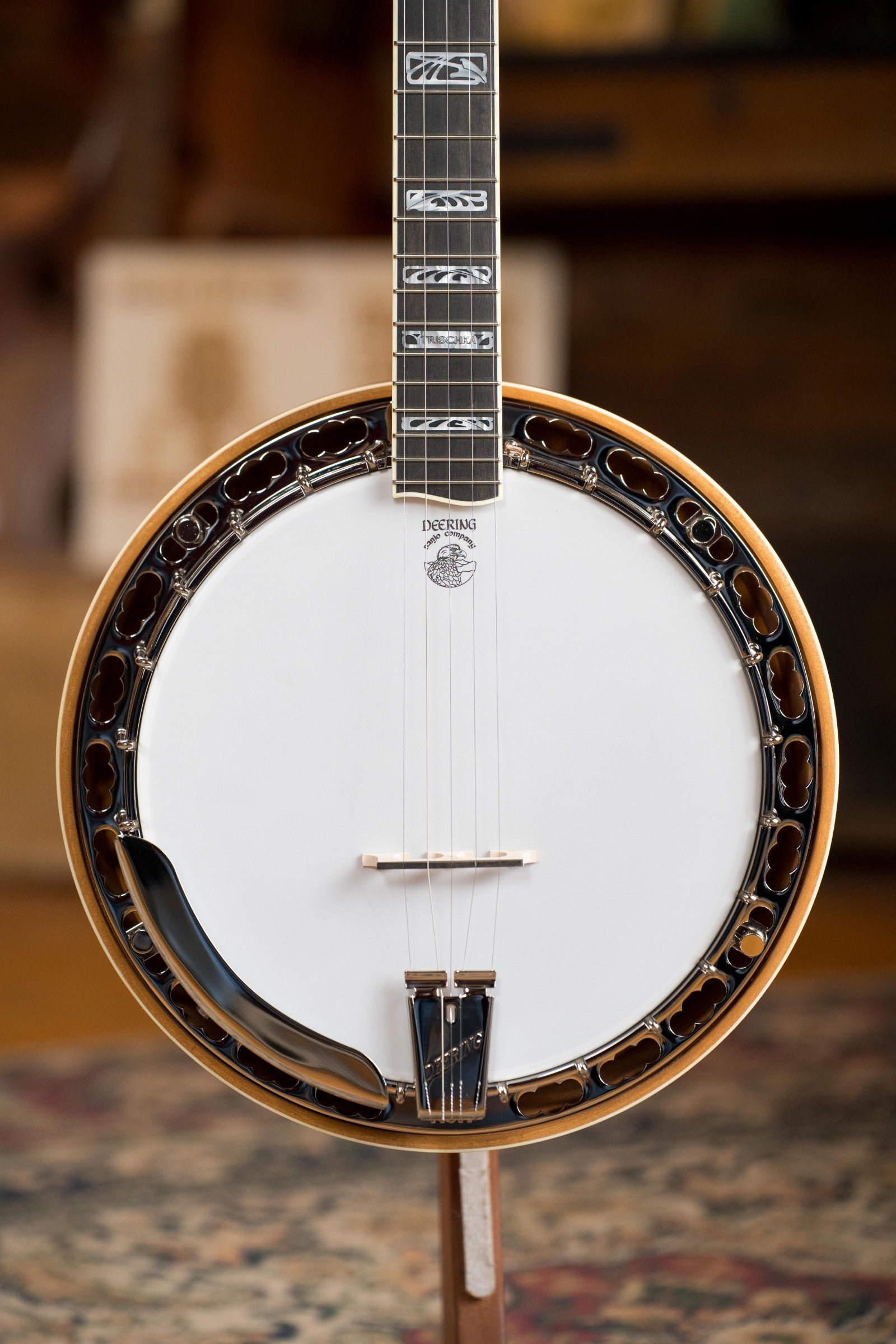 A close-up of the Deering Tony Trischka Silver Clipper Resonator Banjo by Deering Banjos, featuring a white drumhead, metal rim, detailed neck inlays, wood body, and spikes at 7–10—set against a patterned rug for classic bluegrass vibes.