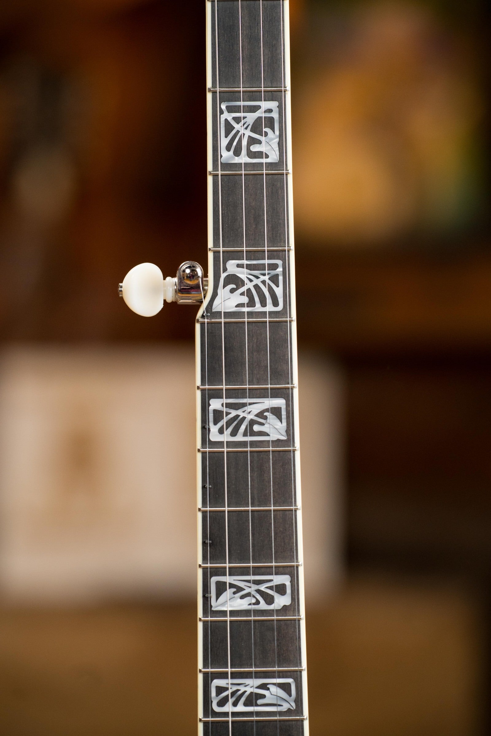 Close-up of a Deering Banjos Tony Trischka Silver Clipper Resonator Banjo w/Spikes at 7, 8, 9, and 10 (#0100), featuring ornate mother-of-pearl fretboard inlays and tuning peg against a blurred brown backdrop—ideal for bluegrass fans.