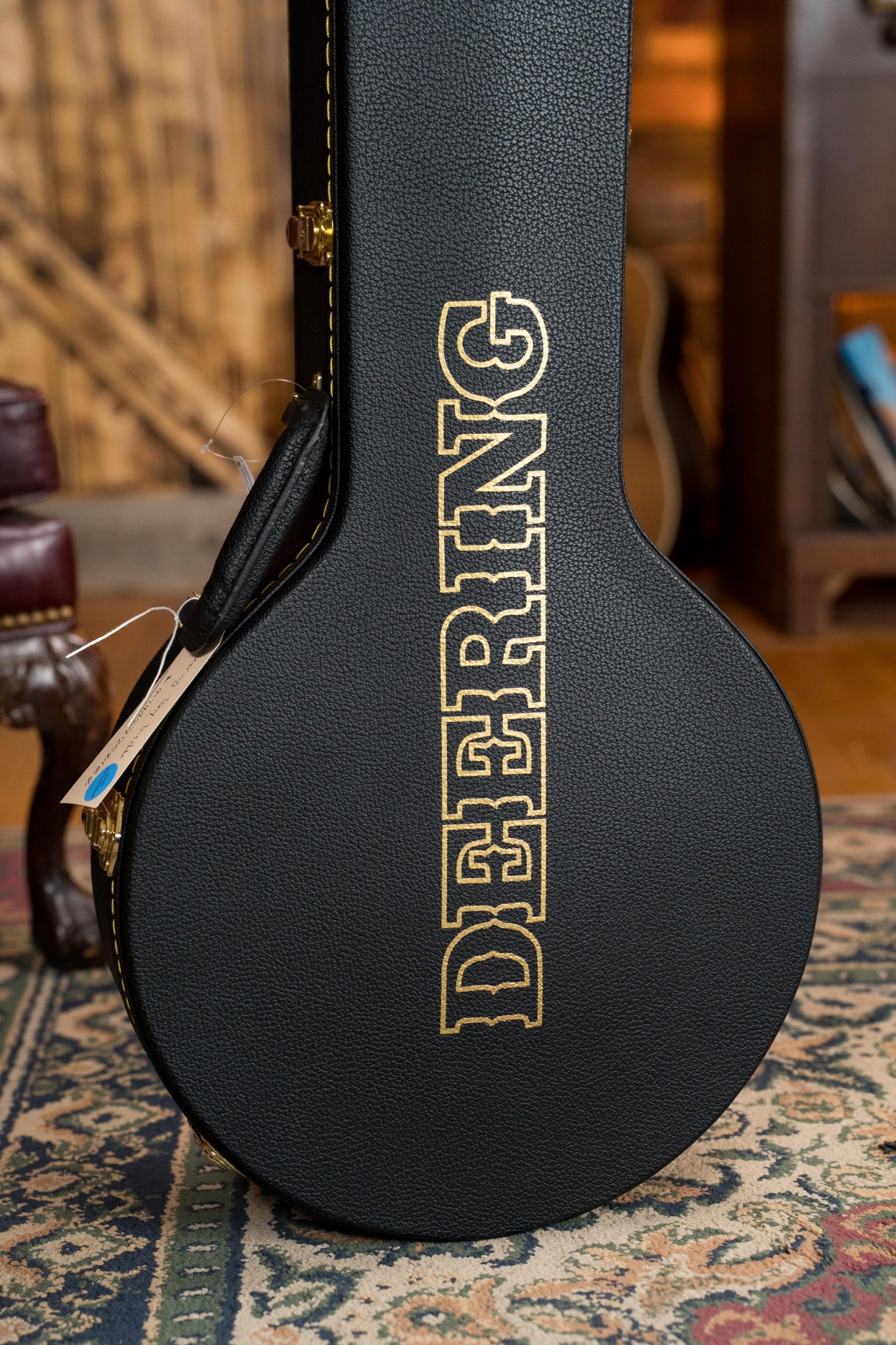 A black Deering Banjos case with gold lettering stands upright on a patterned rug, ready to protect your Tony Trischka Silver Clipper Resonator Banjo w/Spikes Installed at 7, 8, 9, and 10 (#0100) in a warm indoor setting.