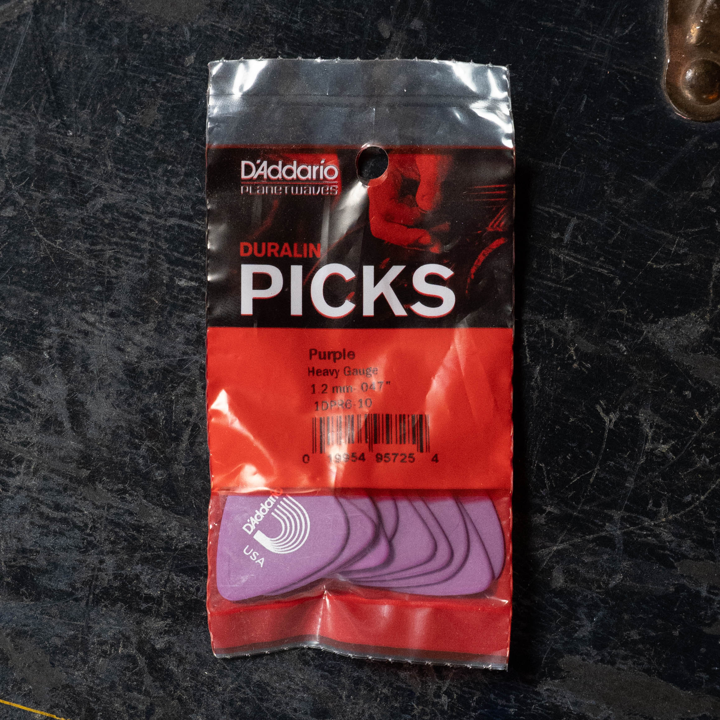 A pack of D'Addario Duralin Flatpicks 10 pk, Heavy Gauge (1.21 mm), sits on a dark textured surface, showing several purple picks through the clear lower half of the package.