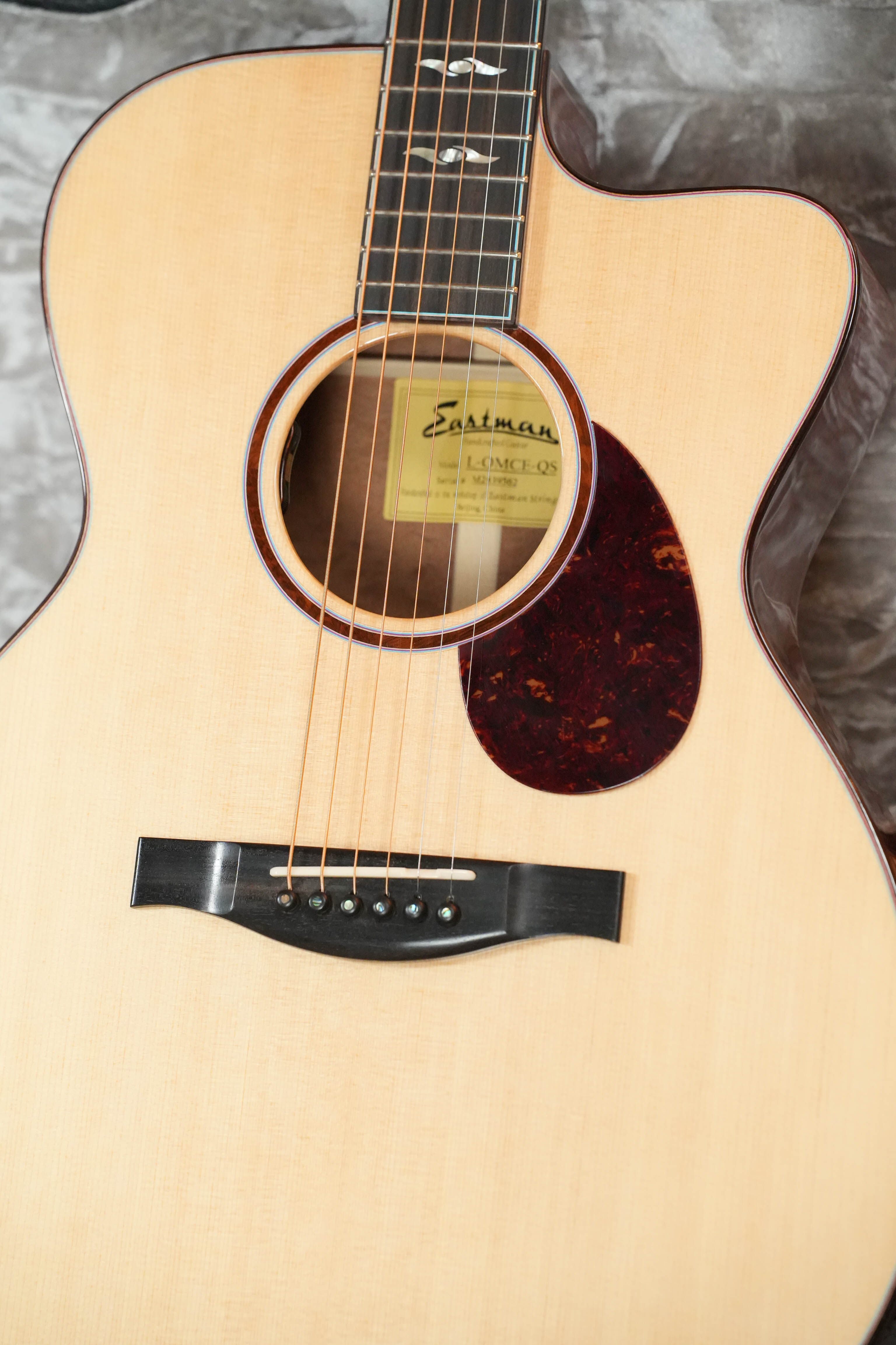 A close-up of an Eastman Guitars *Dread-Not Certified Used* 2024 L-OMCE-QS Orchestra Model acoustic-electric guitar (Engelmann spruce/quilted sapele, LR Baggs VTC, #9562) on a soft gray fabric background.