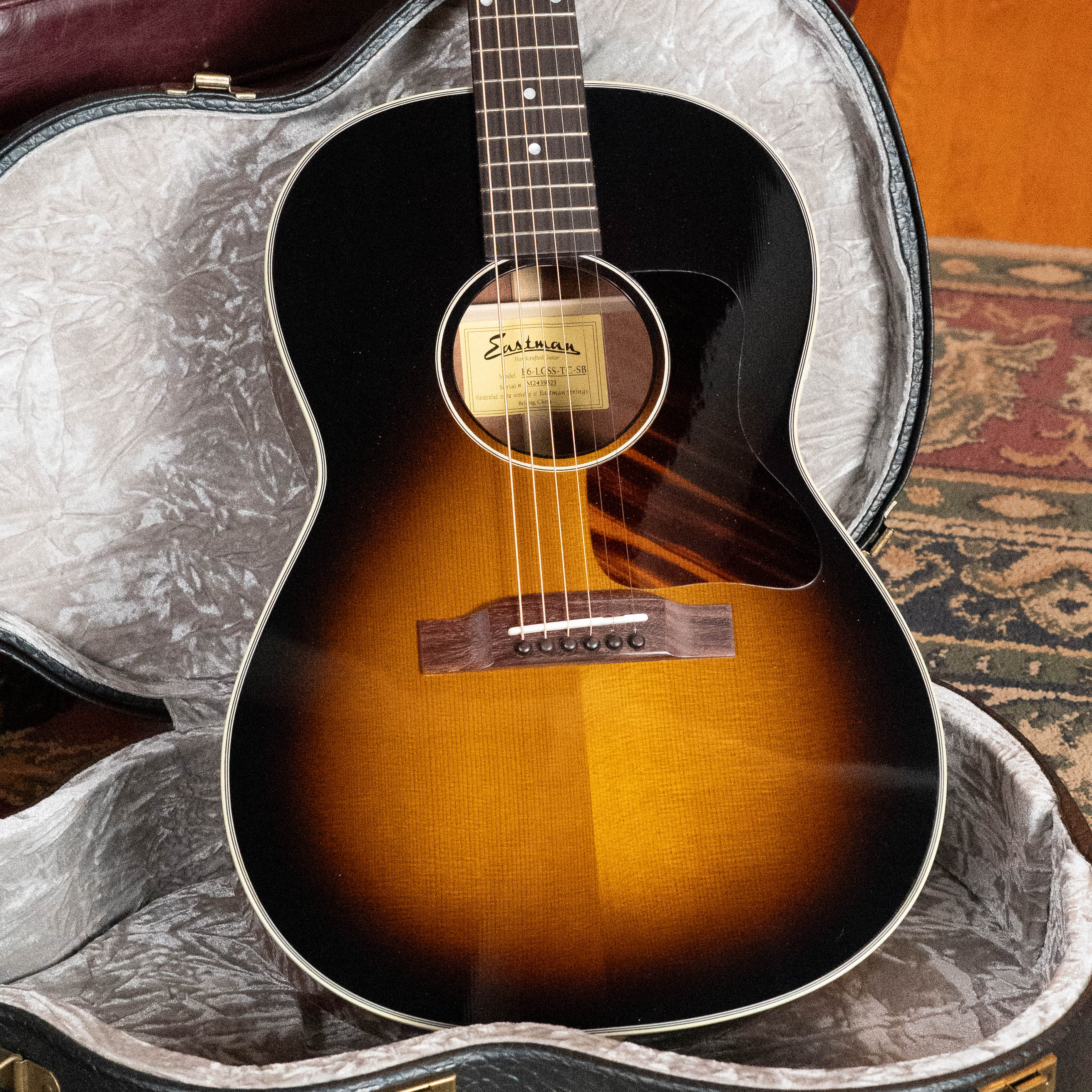 An *Eastman Guitars Dread-Not Certified Used* E6-LGSS-TC-SB Thermo-Cured Sitka/Mahogany LG-Style Slope Shoulder Sunburst Acoustic Guitar (#9323) rests in an open hardshell case on a patterned rug.