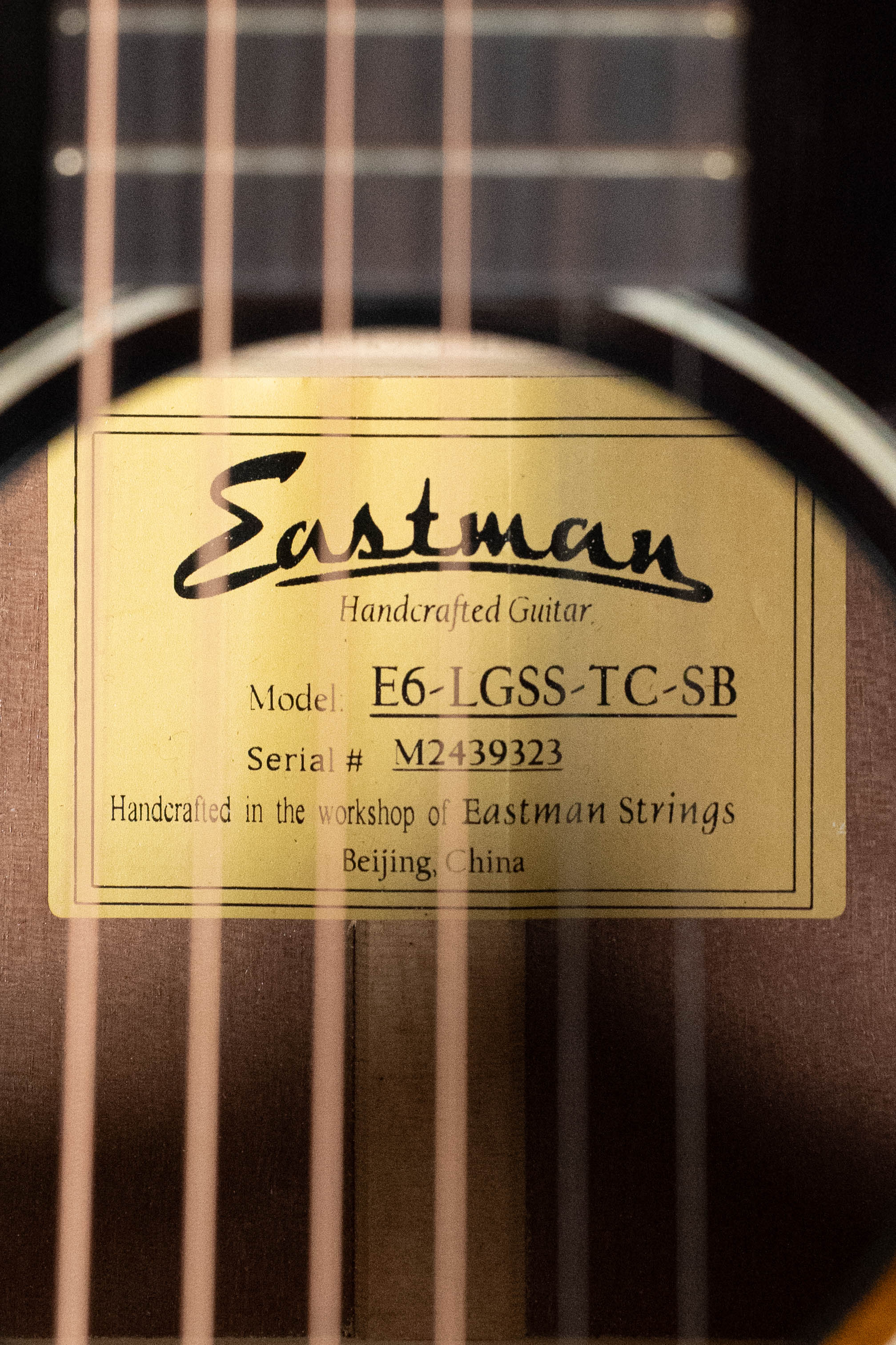 A close-up of the interior label through the sound hole of a *Dread-Not Certified Used* Eastman Guitars E6-LGSS-TC-SB (#9323) Slope Shoulder Acoustic shows details, thermo-cured Sitka top, and workshop info, with strings in the foreground.