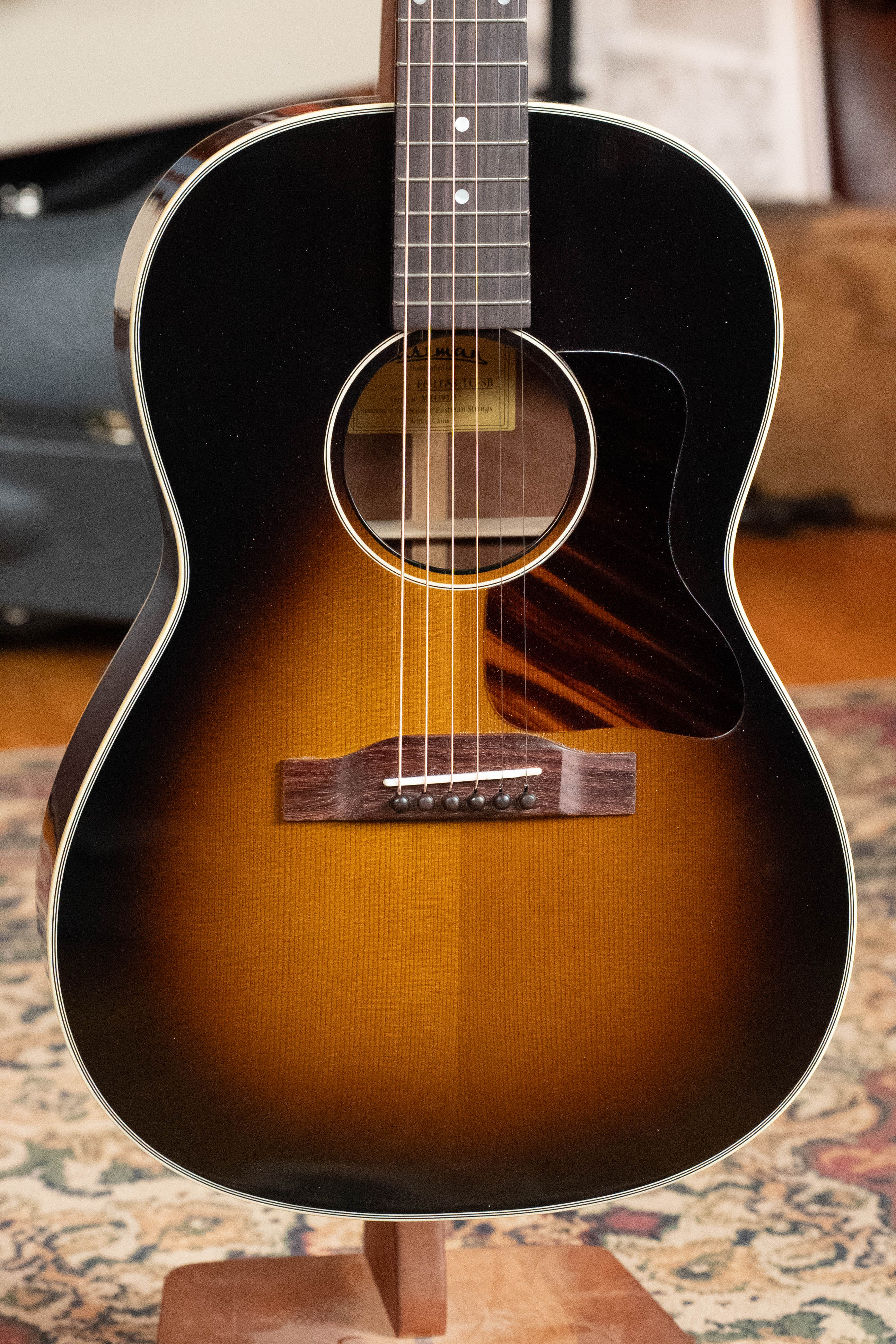 A *Dread-Not Certified Used* Eastman E6-LGSS-TC-SB Thermo-Cured Sitka/Mahogany LG-Style Slope Shoulder Sunburst Acoustic Guitar #9323 by Eastman Guitars rests upright on a stand, with its case in the background atop a patterned rug.