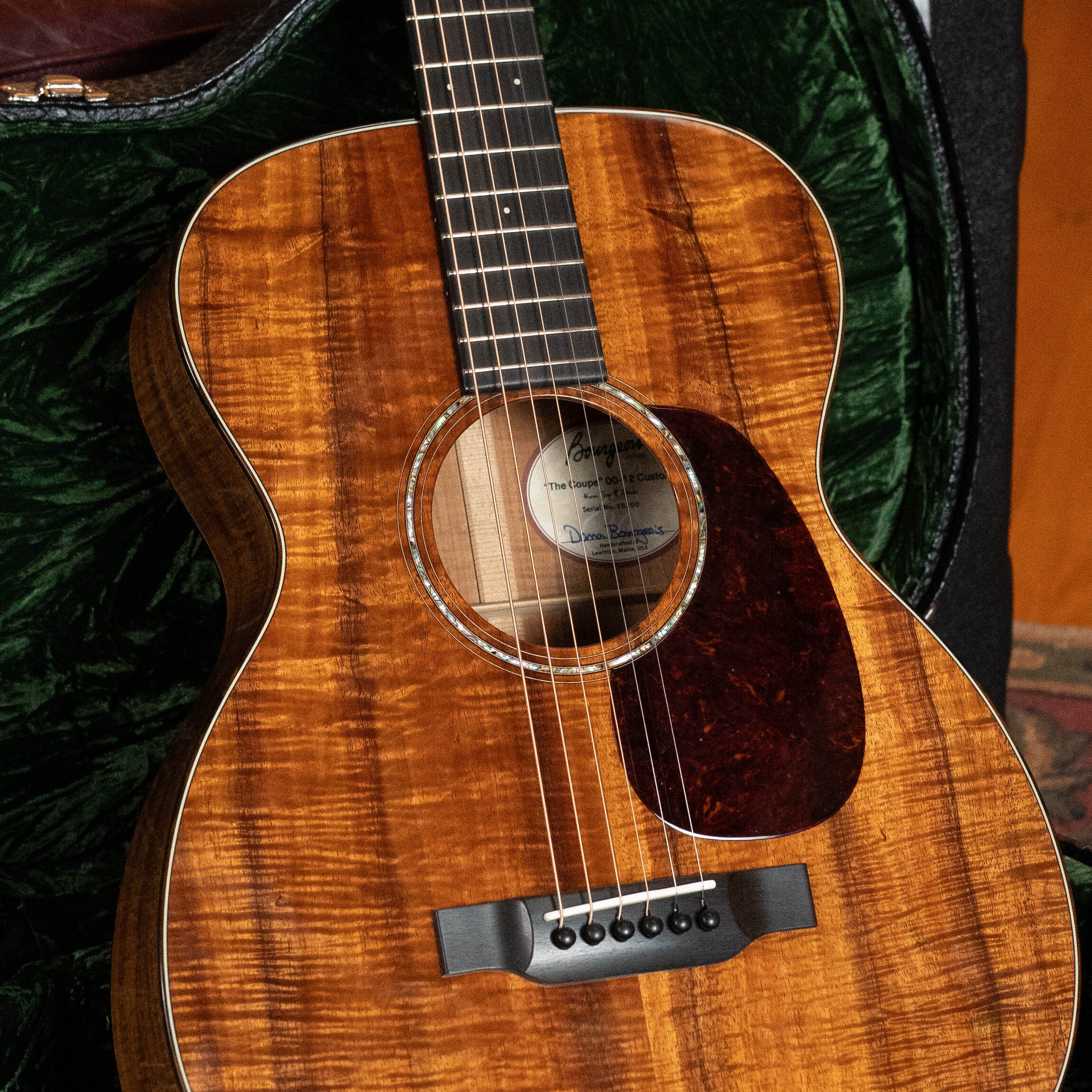 A close-up of a *Dread-Not Certified Used* Bourgeois *Custom Build OO-12 "Coupe" All Figured Koa 12-Fret Acoustic Guitar #0200, featuring a glossy koa finish and dark pickguard, resting in a green plush-lined case.