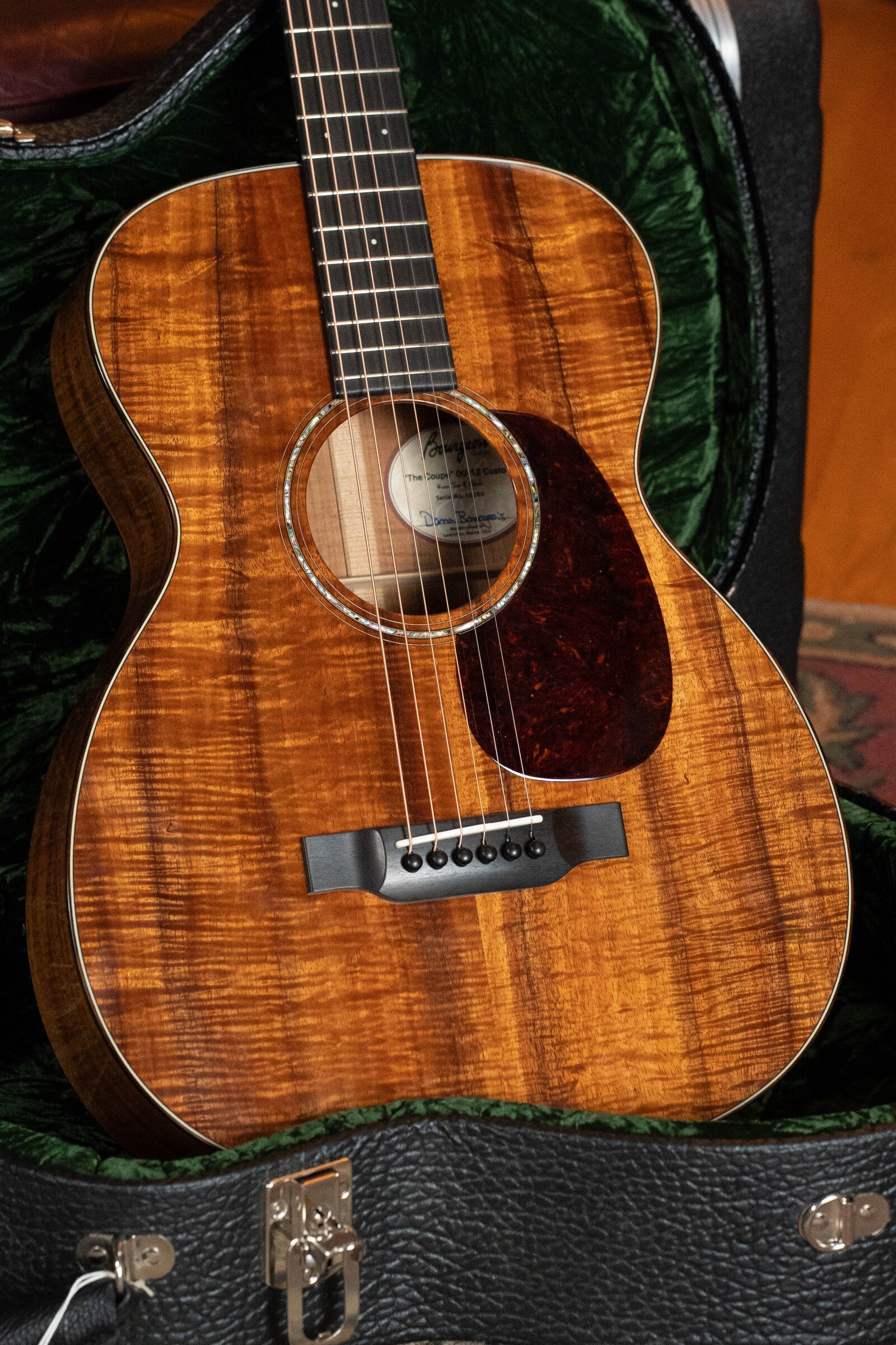 A close-up of a *Dread-Not Certified Used* Bourgeois Custom Build OO-12 "Coupe" All Figured Koa 12-Fret Acoustic Guitar #0200, with a glossy koa body and dark pickguard, shown in its green velvet-lined hard case.