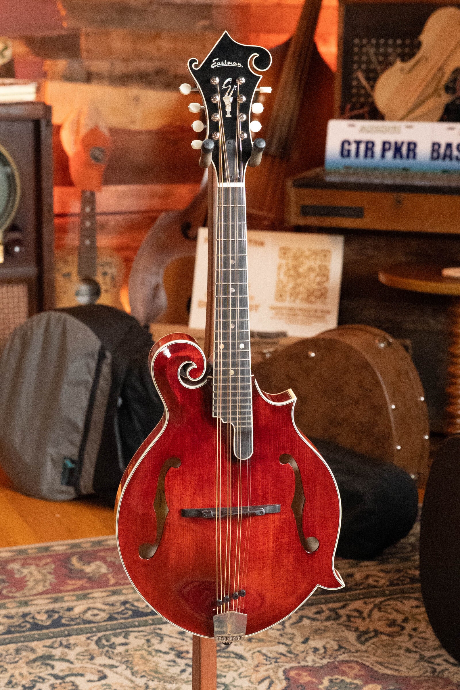 A *Dread-Not Certified Used* Eastman Guitars MDA815 F-Style Mandola Classic Finish #4978 stands on a wooden stand in a cozy room, surrounded by musical instruments and recording equipment.
