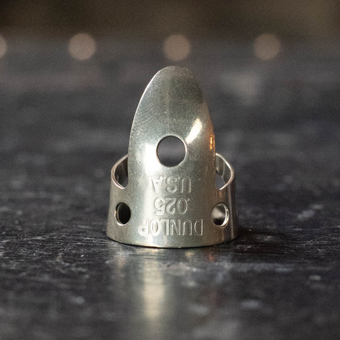 A close-up of the Dunlop Finger Pick .025" (33R025), branded DUNLOP and made of shiny nickel silver, standing upright on a dark textured surfaceβideal for fingerstyle players and banjo enthusiasts.