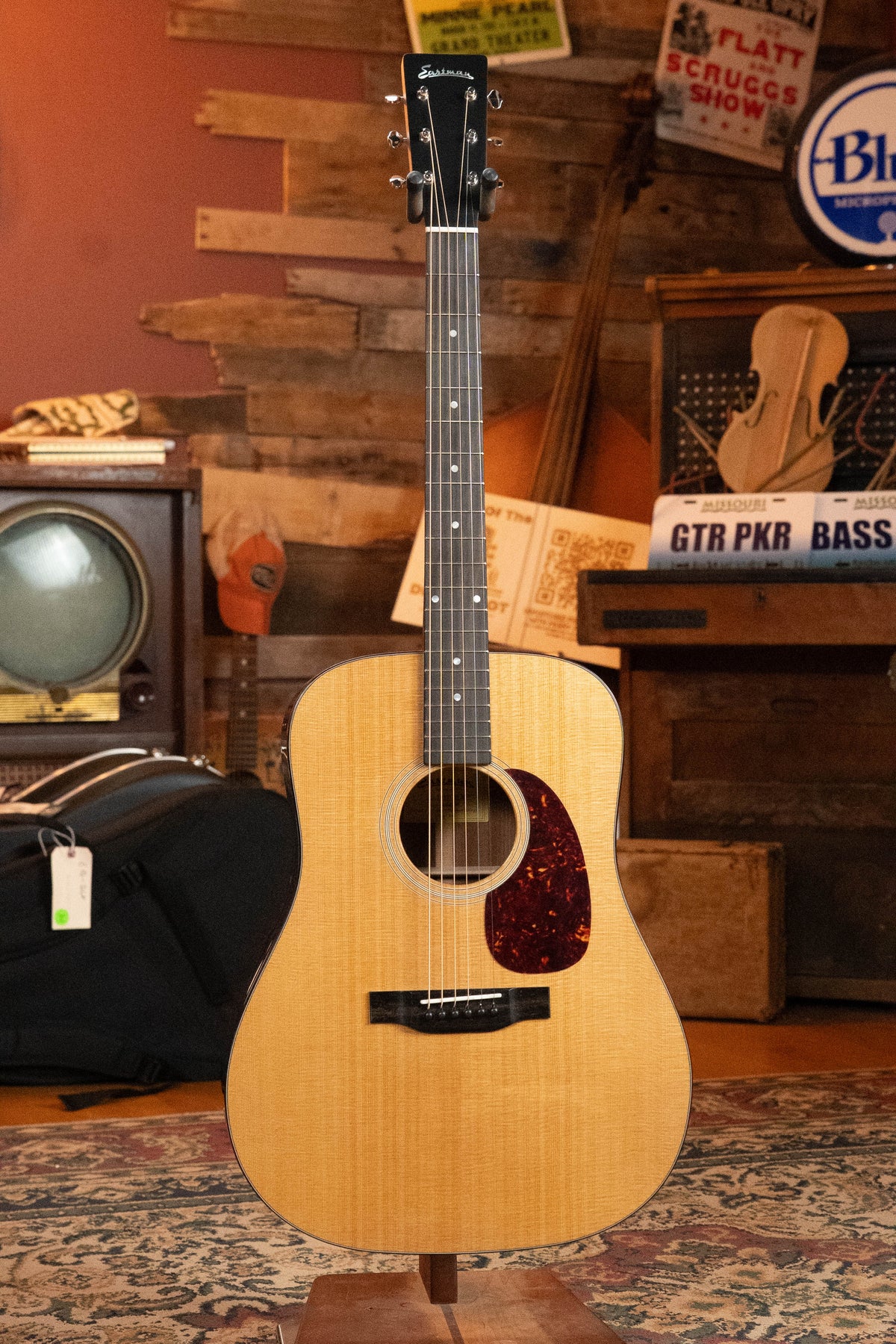 An Eastman Guitars E1D-DLX Sitka/Sapele Dreadnought Guitar w/Fishman Pickup (#6444) stands on a stand in a cozy, vintage-style room with rustic decor, musical gear, a dark headstock, and a tortoiseshell pickguard.
