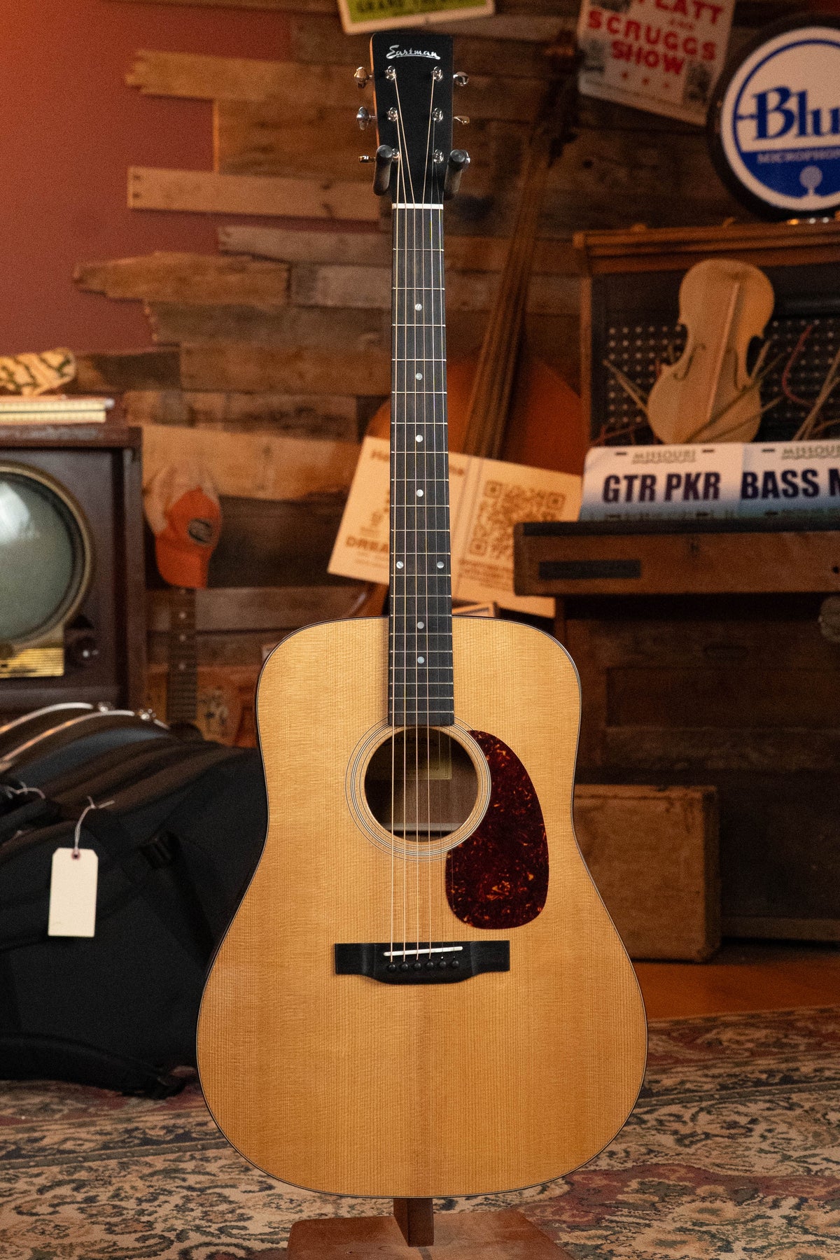 The Eastman Guitars E1D-DLX Sitka/Sapele Dreadnought Guitar w/Fishman Pickup (#7728) stands upright on a display in a cozy, rustic room decorated with musical instruments and vintage items.