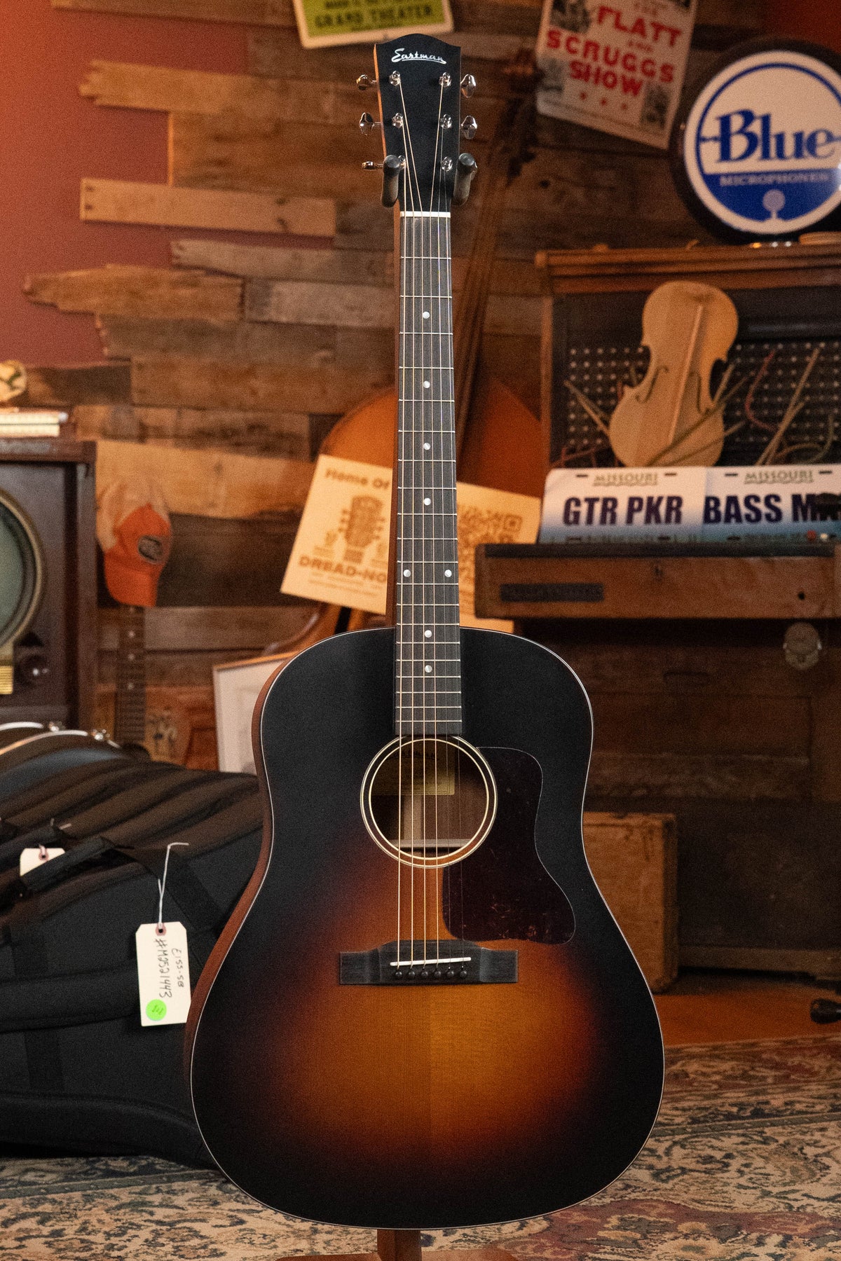 The Eastman Guitars E1SS-SB Sitka/Sapele Sunburst Slope Shoulder Dreadnought Acoustic Guitar #8068 stands on a rug in a cozy, vintage-style room with wood-paneled walls, music gear, and music-themed signs in the background.