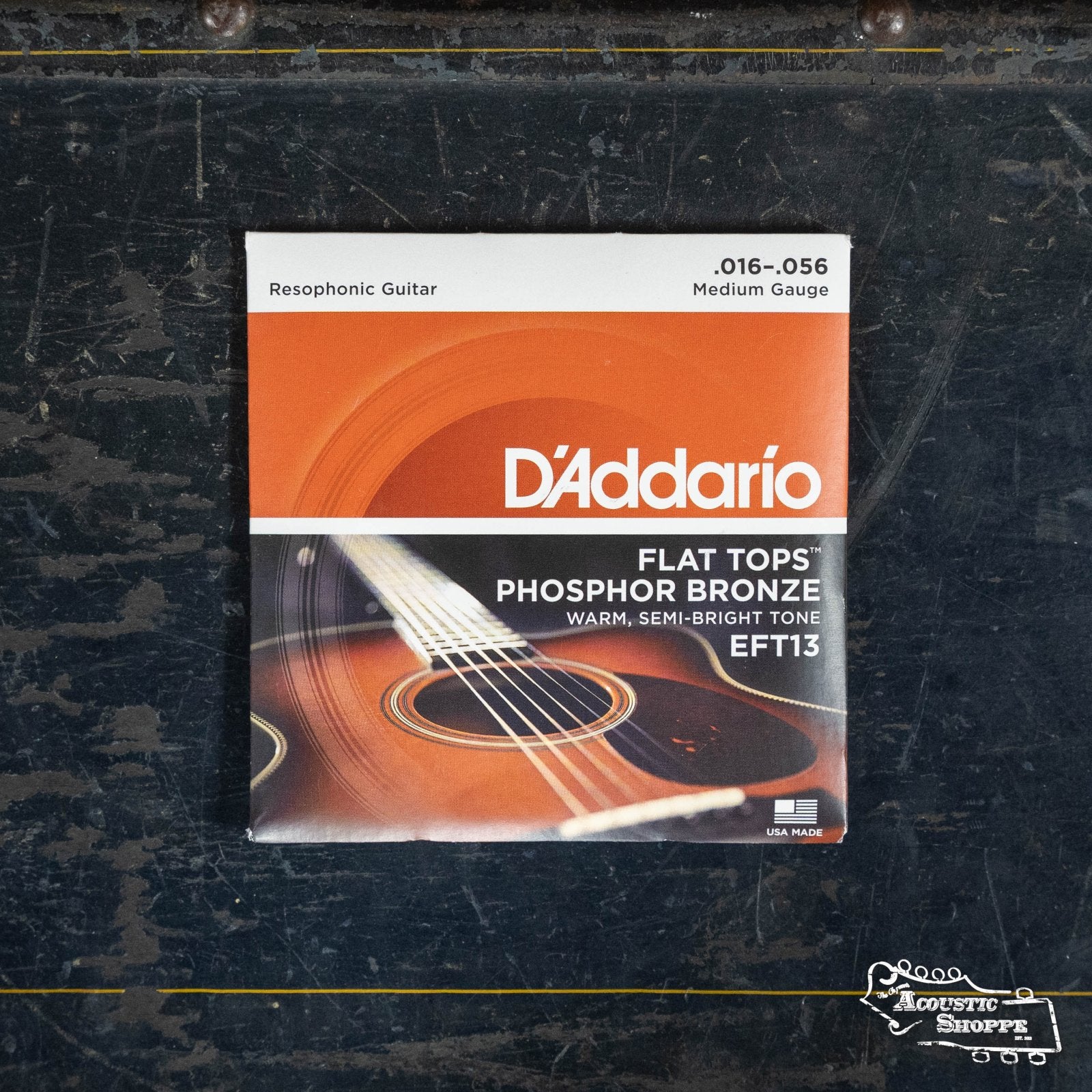 A pack of D'Addario EFT13 Medium Resophonic Flat Tops Phosphor Bronze Acoustic Guitar Strings (16-56) sits on a dark, textured surface. The packaging, branded D'Addario, features a guitar image.