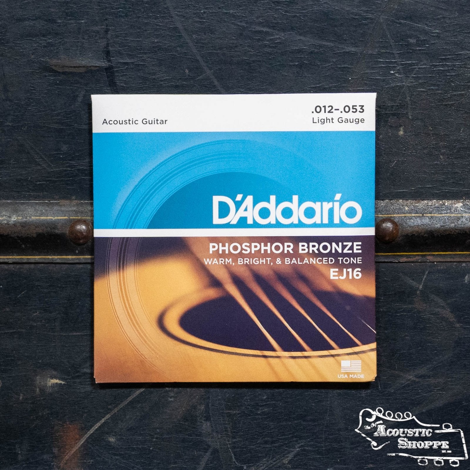 A pack of DAddario EJ16 Phosphor Bronze Acoustic Guitar Strings, Light 12-53, featuring long-lasting tone. Shown with a close-up of the strings on the package, set against a dark, textured surface.