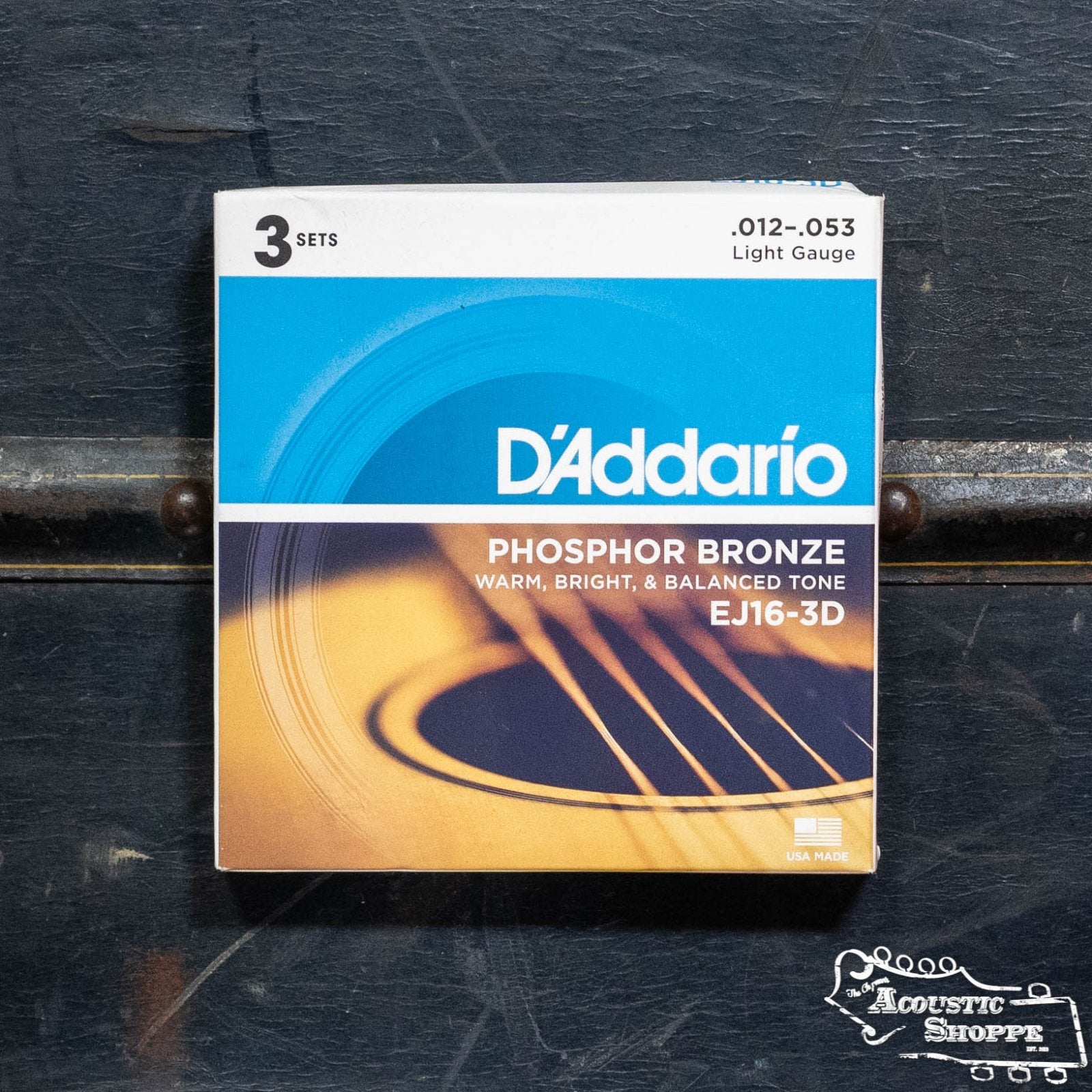 A box of D'Addario EJ16-3D Phosphor Bronze Acoustic Guitar Strings 3 Set Light 12-53 sits on a dark, textured surface with a partial guitar graphic and the Acoustic Shoppe logo, noted for their long-lasting tone.