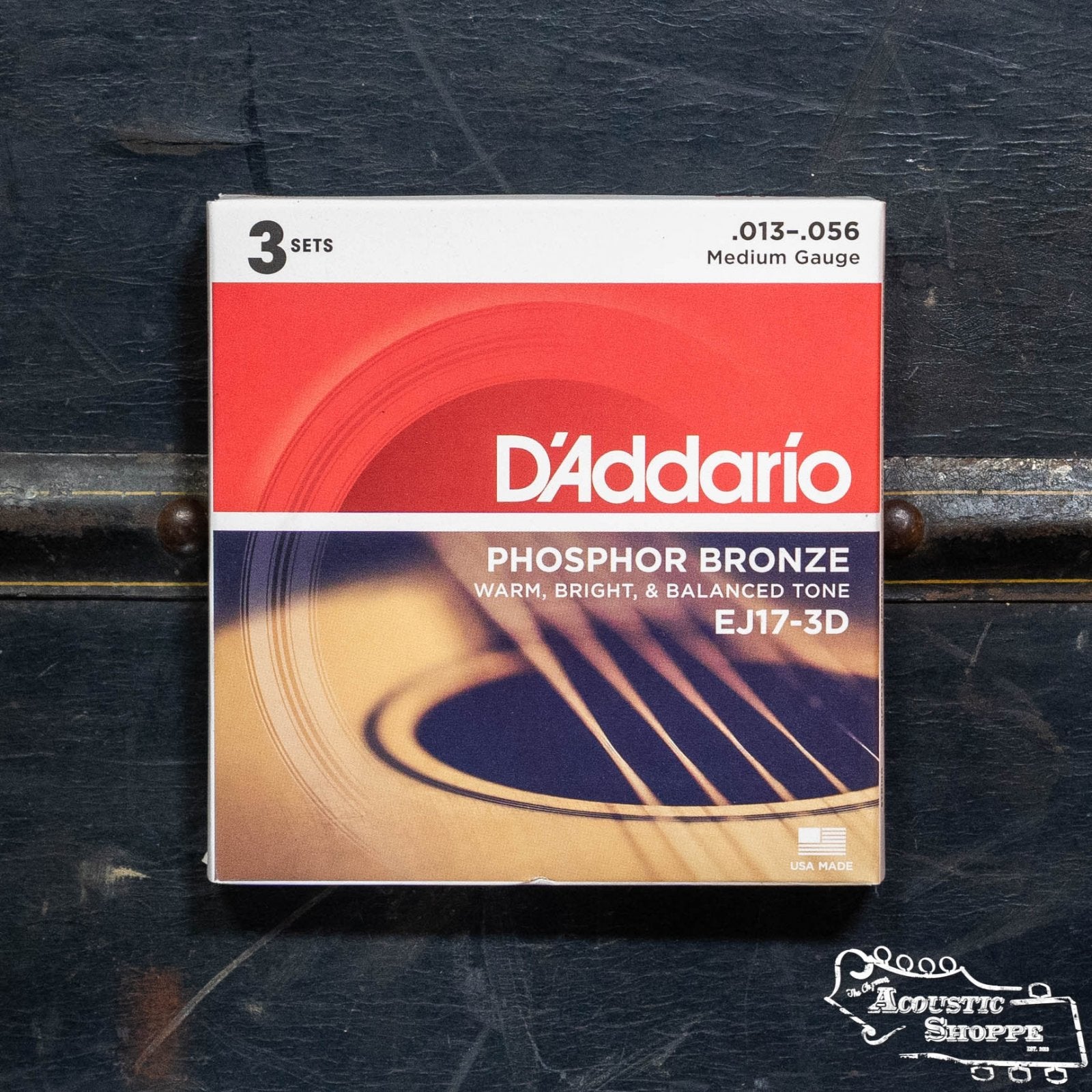 A box of D'Addario EJ17-3D Phosphor Bronze Acoustic Guitar Strings, Medium 13-56 (3 sets), with red, orange, and white packaging, rests on a dark surface—designed for rich acoustic tone and stamped with the Acoustic Shoppe logo.