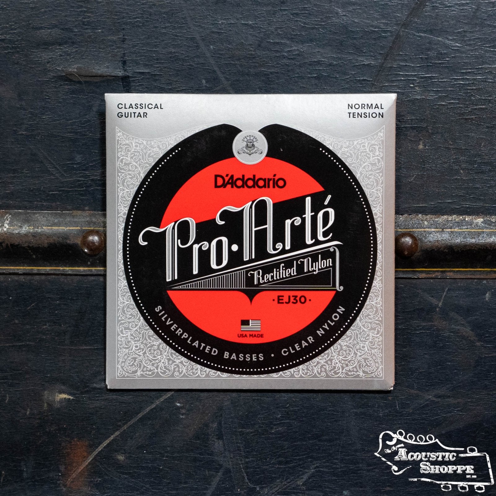 A pack of DAddario EJ30 Pro-Arte Rectified Trebles Normal Tension Acoustic Guitar Strings rests on a dark surface, featuring black, red, and silver packaging with ornate details and the DAddario branding.