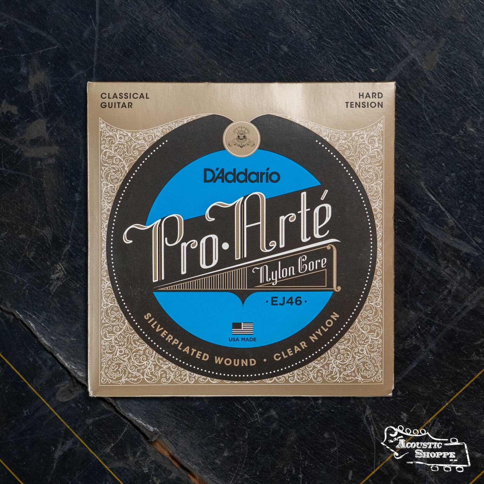 A pack of DAddario EJ46 Pro-Arte Nylon Classical Guitar Strings (Hard Tension) with a gold, brown, and blue design rests on a dark, textured surface.