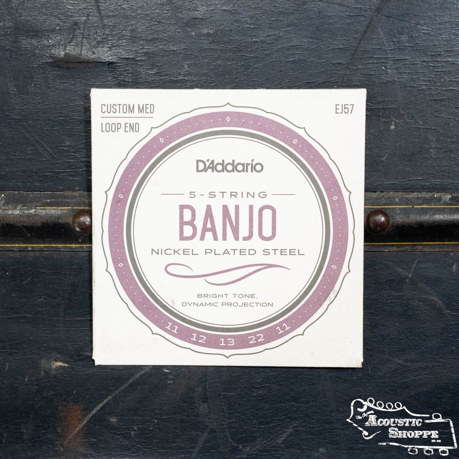 A packet of DAddario EJ57 Banjo Nickel Custom Medium 11-22 strings for 5-string banjos, crafted from nickel plated steel, is shown on a dark surface with a round label featuring “Bright Tone, Dynamic Projection” and The Acoustic Shoppe logo.