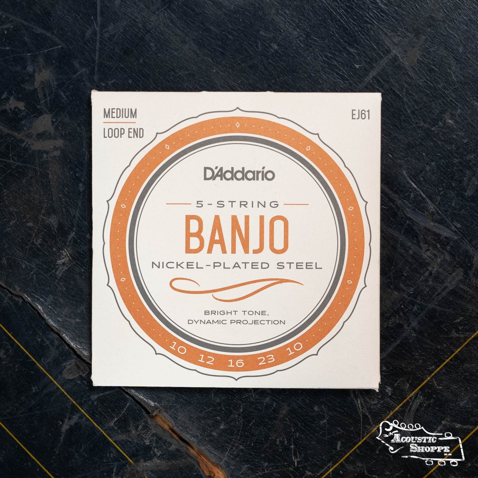 A pack of DAddario EJ61 5-String Banjo Strings, Nickel, Medium 10-23, sits on a dark textured surface. The label displays medium gauge and loop end—essential acoustic accessories for banjo players.