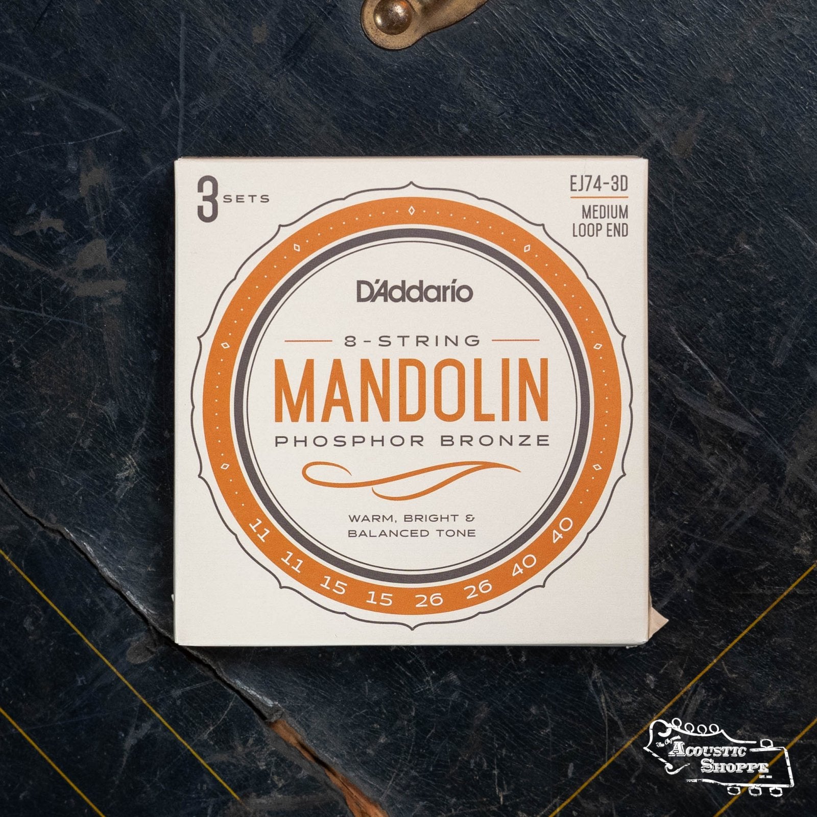 A box of DAddario EJ74-3D Mandolin Strings, Phosphor Bronze, Medium (11-40), 3 sets, sits on a dark, worn wooden surface. The white packaging with orange accents shows gauges and bold DAddario branding.