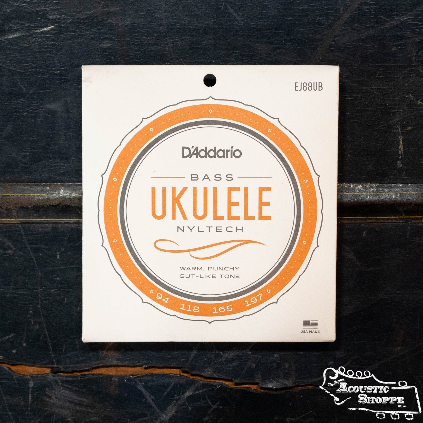 A pack of D'Addario EJ88UB Nyltech Ukulele Strings Bass sits on a dark wooden surface. The white, orange, and gray packaging highlights their warm, punchy, gut-like tone for bass ukuleles.