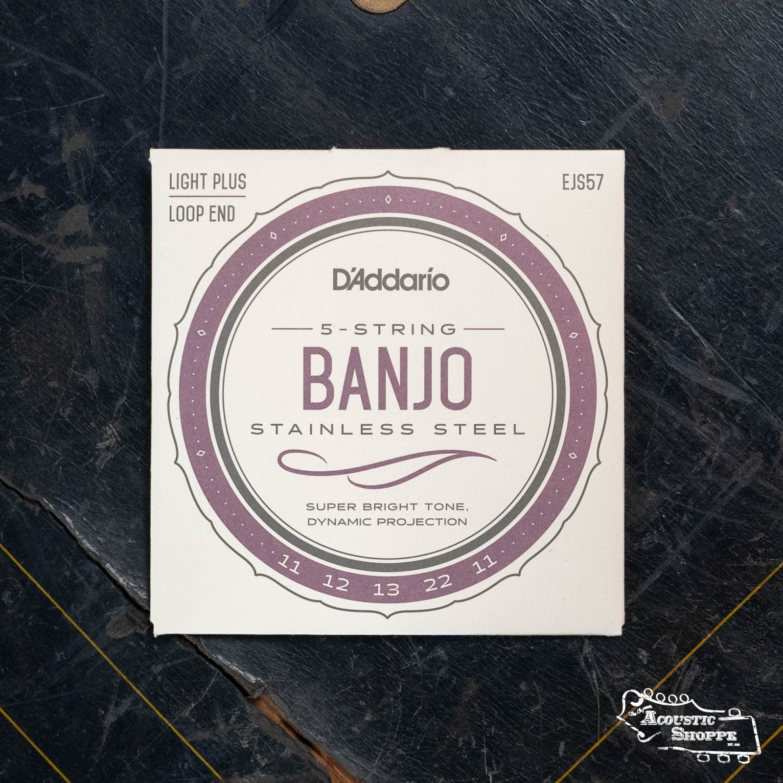 A pack of D’Addario EJS57 5-String Banjo Strings, Stainless Steel, Custom Medium (11-22), is shown on a dark surface. The packaging mentions Super Bright Tone and Dynamic Projection—ideal for heavy picking.
