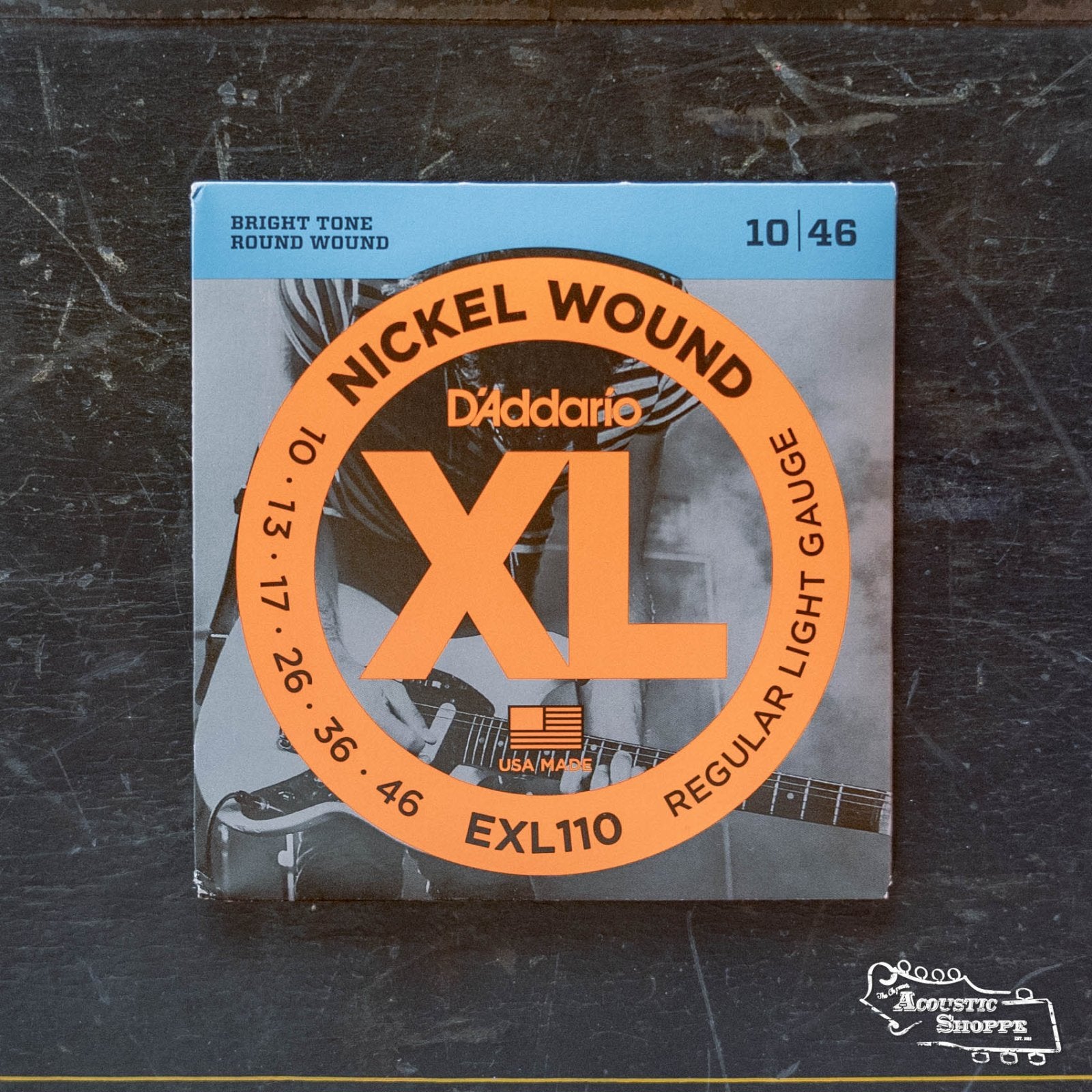 A pack of D'Addario EXL110 Nickel Wound Electric Guitar Strings, Regular Light, 10-46, sits on a dark surface. The packaging displays the D'Addario name, an orange circle, and a grayscale guitar image in the background.