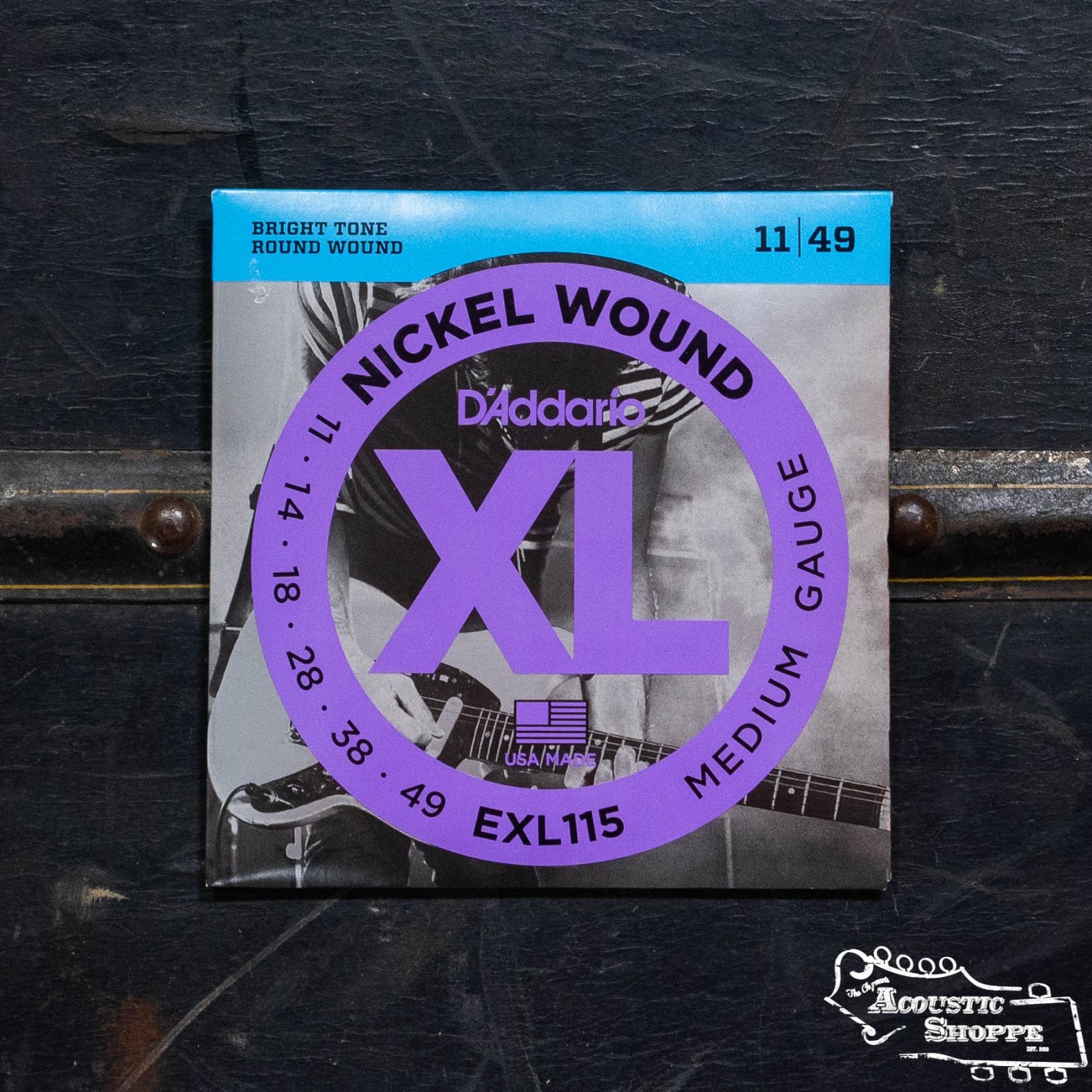 A pack of D'Addario EXL115 Nickel Wound Electric Guitar Strings Medium 11-49 sits on a dark surface, featuring a close-up guitarist image and the Acoustic Shoppe logo in the bottom right corner.