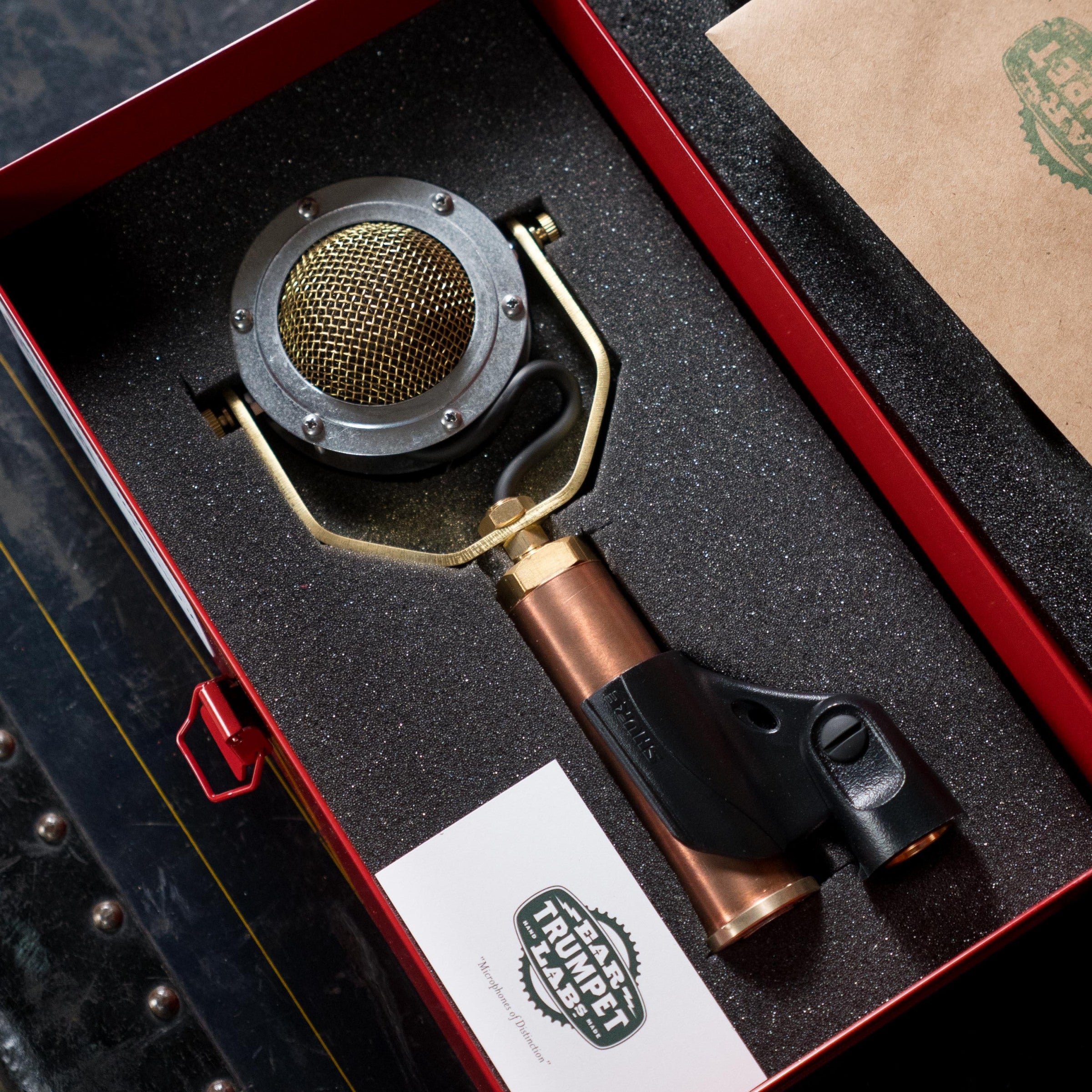The Ear Trumpet Labs Delphina Microphone, a vintage-style large-diaphragm condenser with a gold mesh head and copper body, rests in a foam-lined red box beside an Ear Trumpet Labs business card.